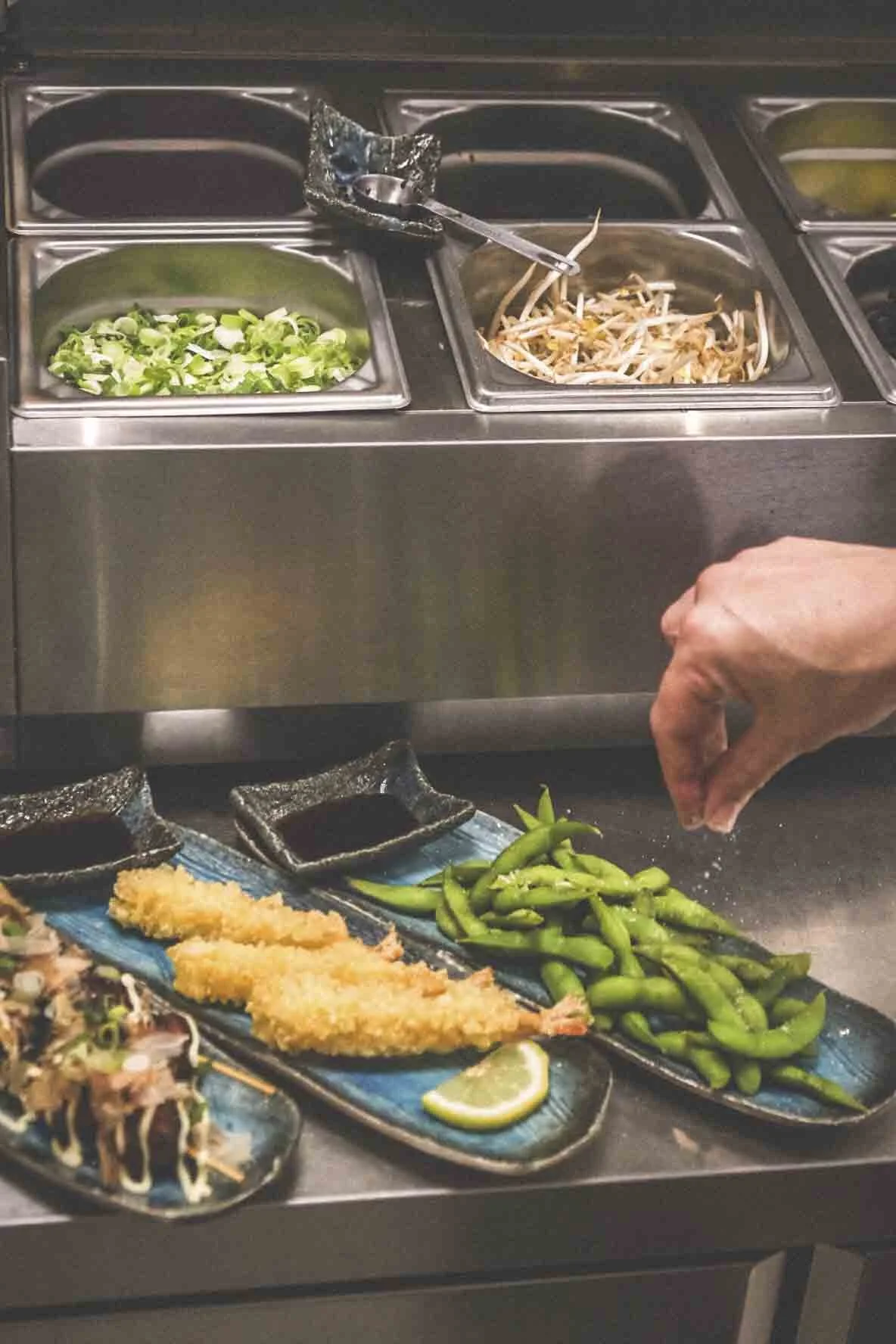 Plat de sushi frit sur une assiette bleue avec une tranche de citron vert et des haricots verts à côté, un comptoir avec des légumes frais et des condiments, une main saupoudrant probablement du sel ou du poivre.