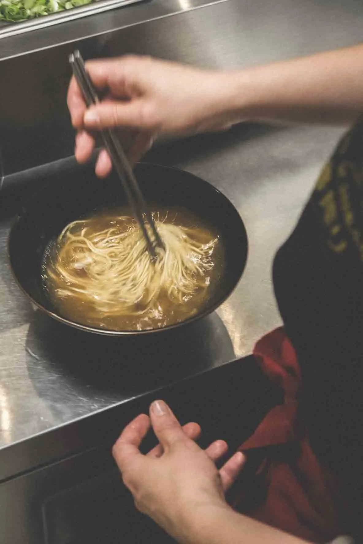Une personne utilise des baguettes pour mélanger des noodles dans un bol noir, posée sur une table en métal.
