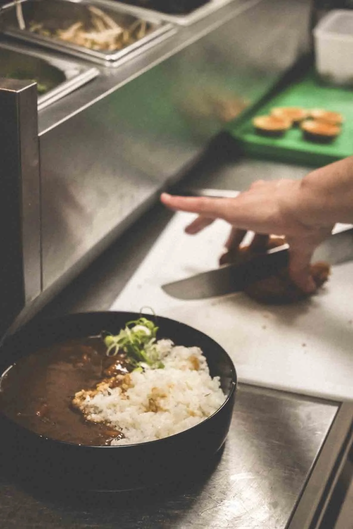 Une assiette de riz blanc avec un curry ou sauce brune et des garnitures vertes, placé sur une surface de cuisine en acier inoxydable, avec une main qui coupe un aliment sur une planche verte en arrière-plan.
