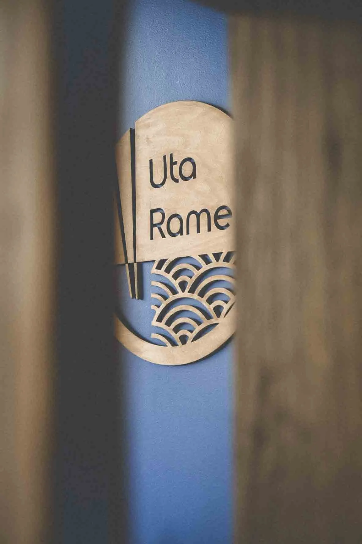 Un panneau en bois avec l'inscription "Uta Rame" et un motif de vagues stylisées en bas, fixé à un mur bleu.