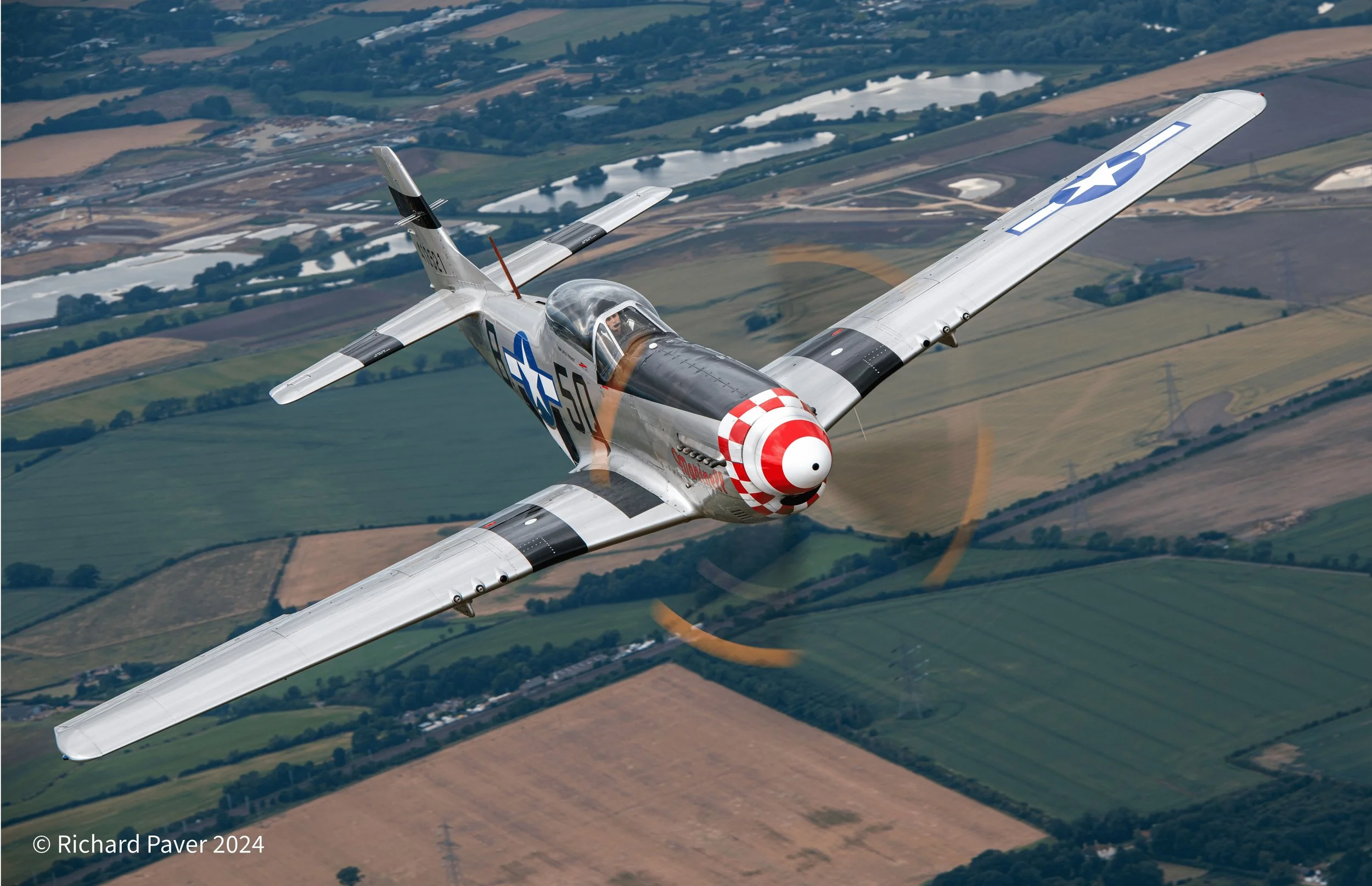 A vintage military jet airplane flying over farmland with patchwork fields, bodies of water, and trees below.