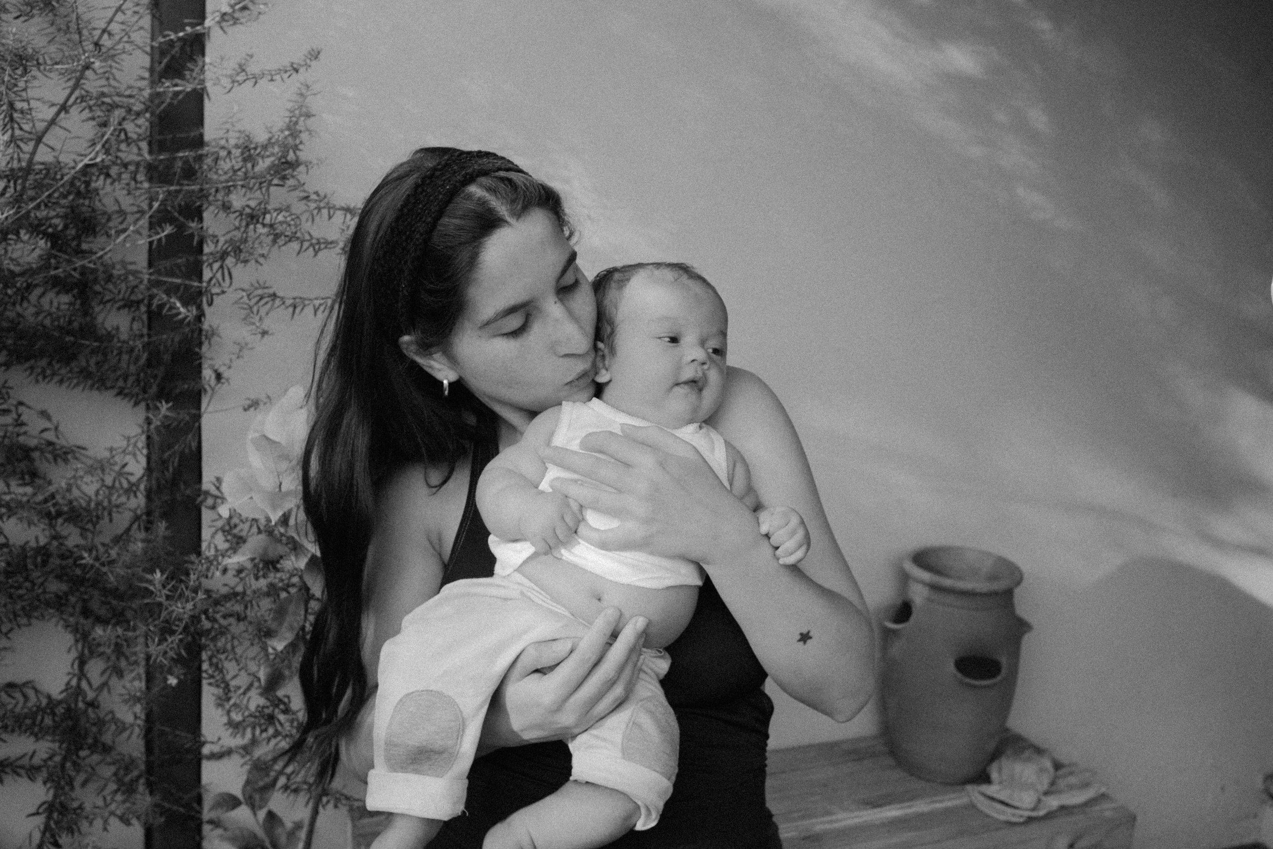 A woman with dark hair and a headband holding a baby with light hair in a room. The woman is gently kissing the baby's head. There are plants and a star-patterned pot in the background.