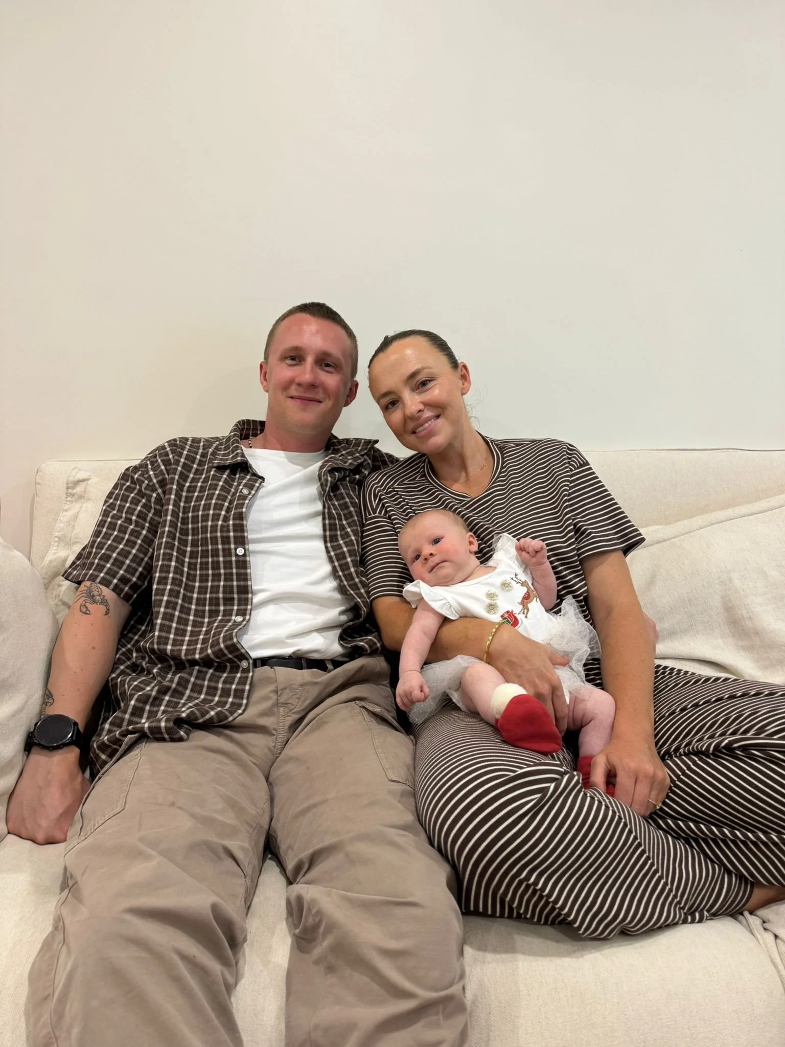 A family sitting on a beige couch against a plain white wall, consisting of a man, woman, and baby. The man is wearing a brown checkered shirt and beige pants, the woman is dressed in a striped brown and white shirt and matching pants, and the baby is dressed in a white outfit with Christmas-themed embroidery, red socks, and is being held by the woman.