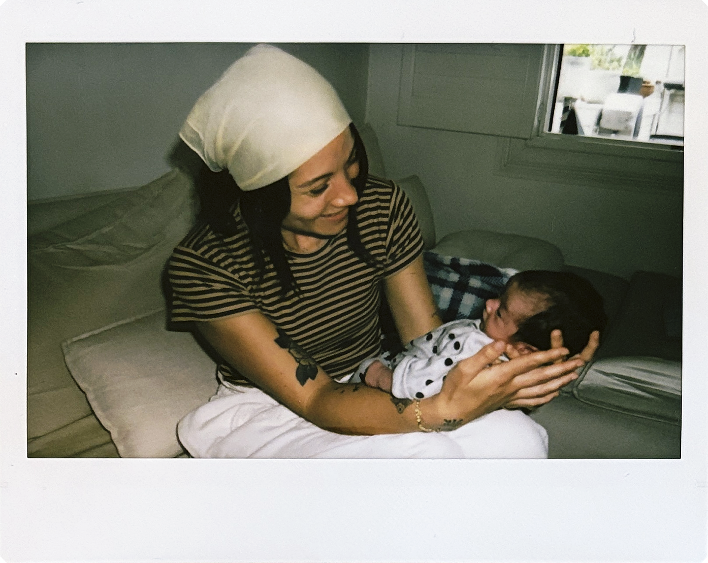 A woman with dark hair, wearing a headscarf and a black and white striped shirt, is smiling and holding a baby.
