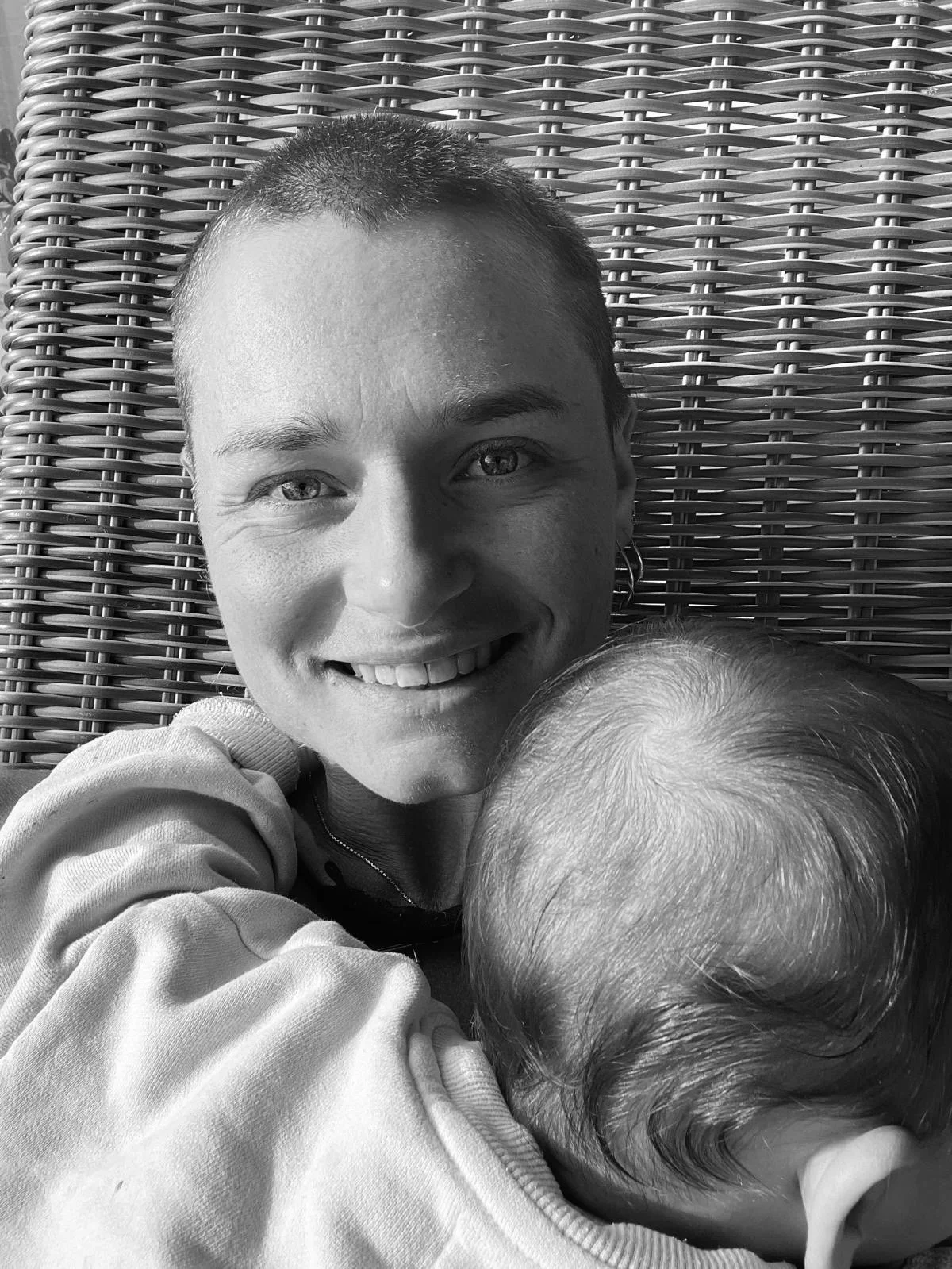 A smiling person with short hair lying on a woven surface, holding a young child with short hair close to her face.