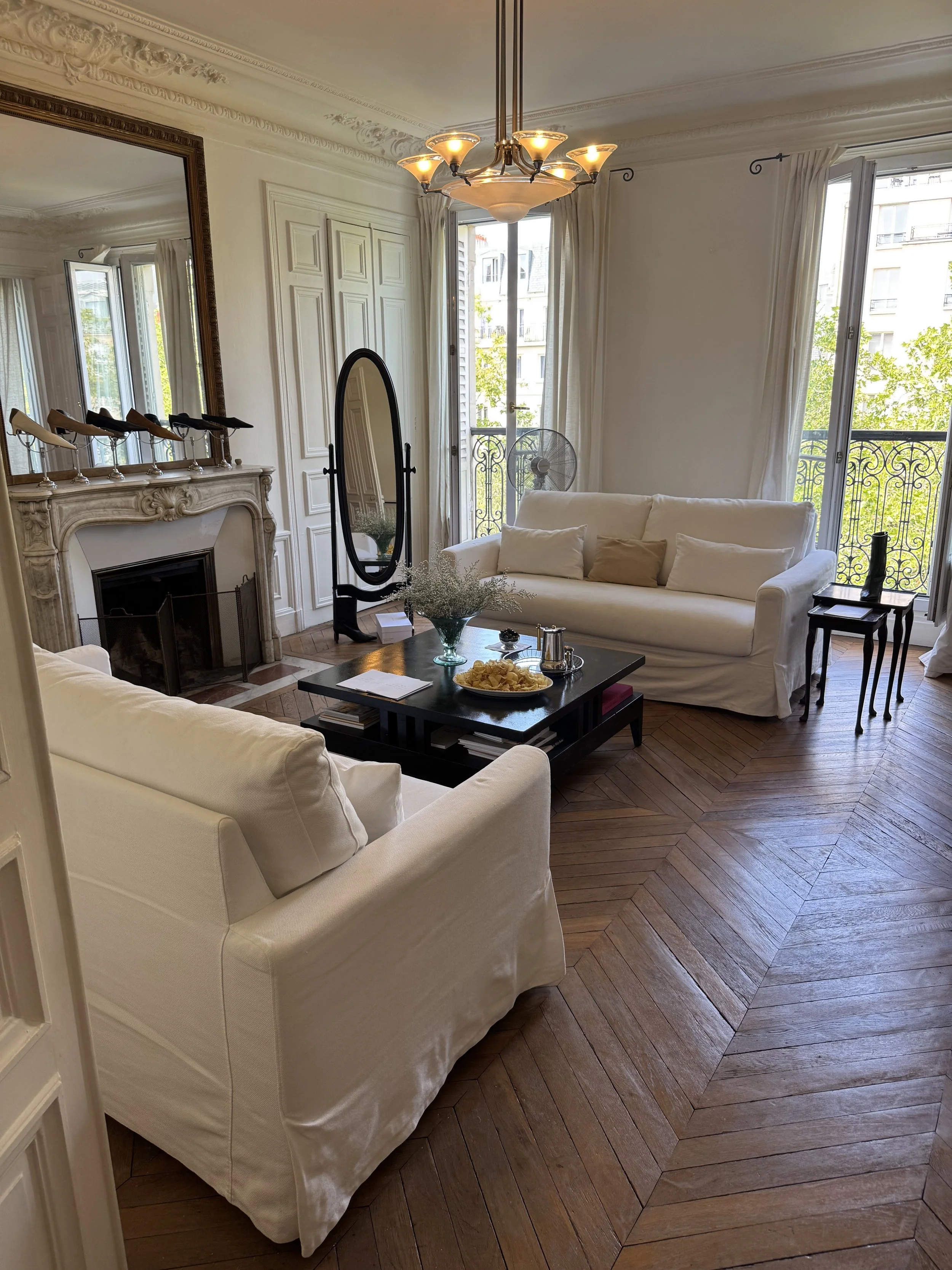 Living room with white sofas, a fireplace with a decorative mantle, a large mirror, a standing mirror, a fan, and large windows with white curtains showing outdoor trees and buildings, hardwood floors.