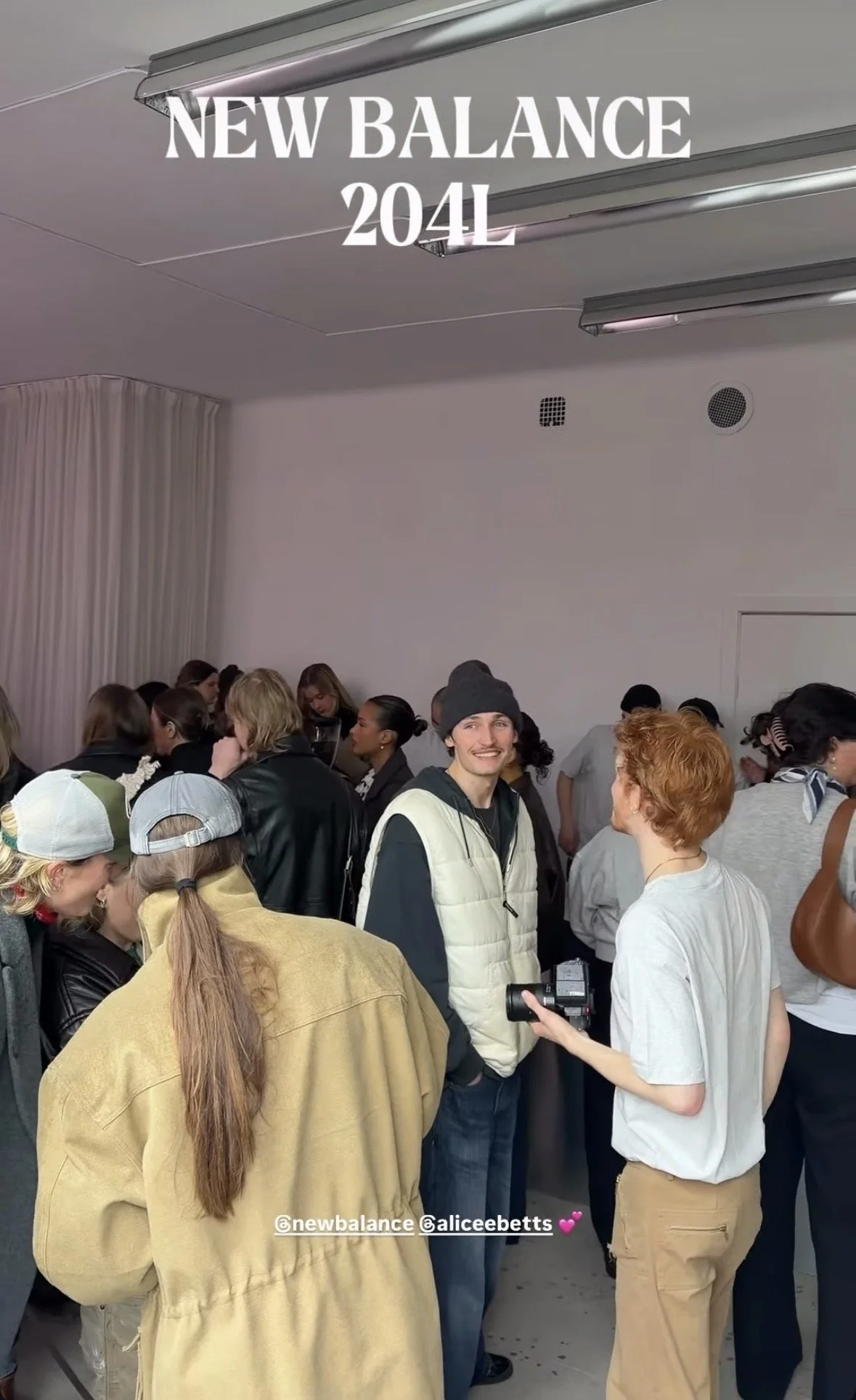 Group of people gathered at a New Balance event, with some members engaging in conversations, and two individuals in focus—one man with a beanie smiling, and another person holding a camera. The background shows a plain white wall with a ceiling mounted light fixture.