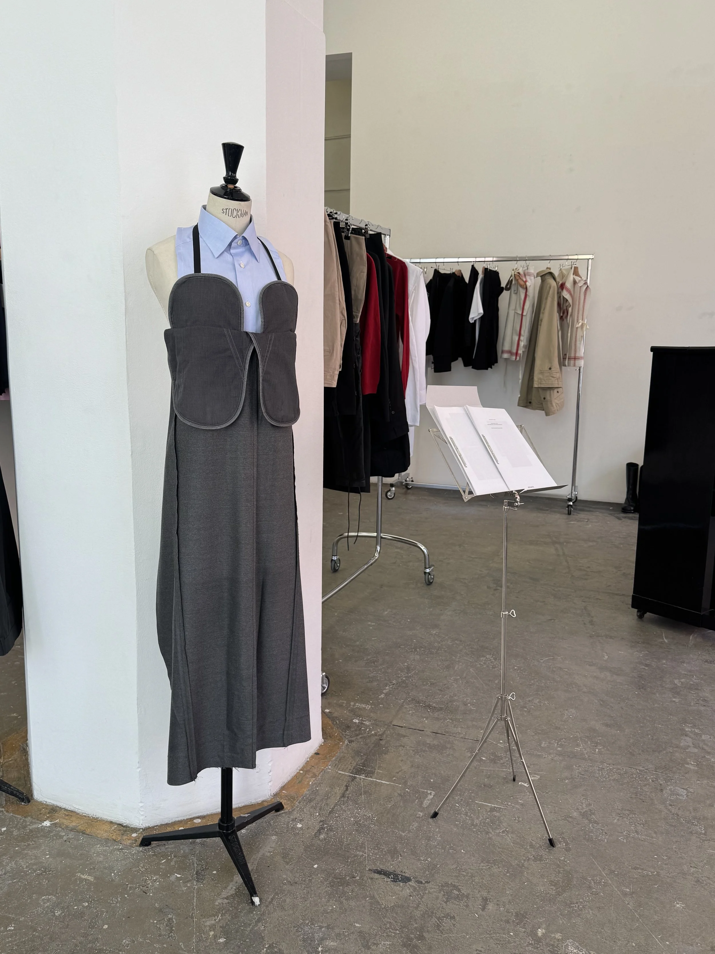 A clothing display with a mannequin dressed in a blue shirt, gray skirt, and an apron, with a clothing rack holding various jackets and shirts in the background, and a stand with papers or brochures.