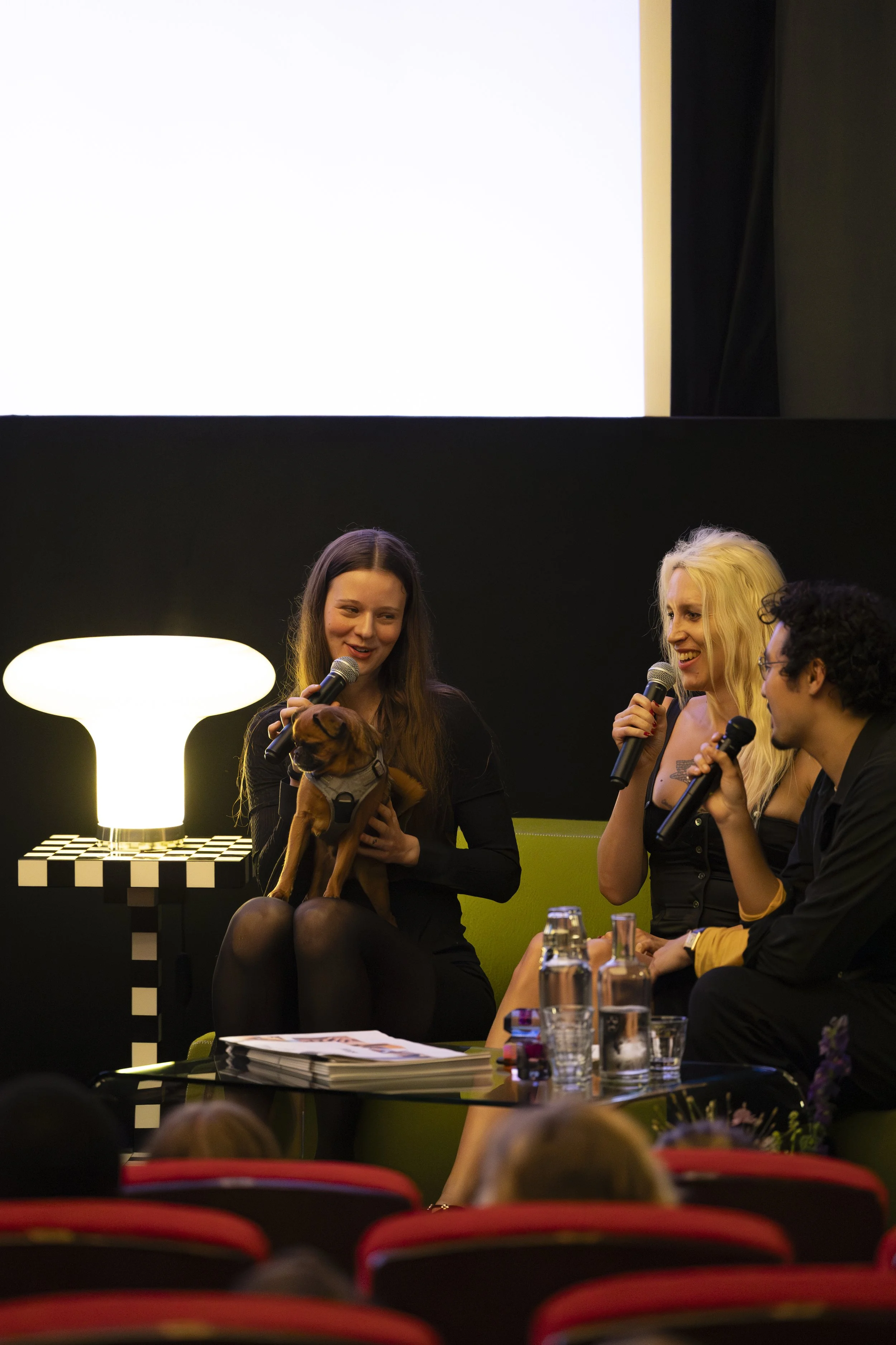 Four people sitting on stage during a panel discussion, with three women holding microphones. One woman is holding a small brown dog. They are seated in front of an audience, with a black background, a white lamp, and a large screen overhead.