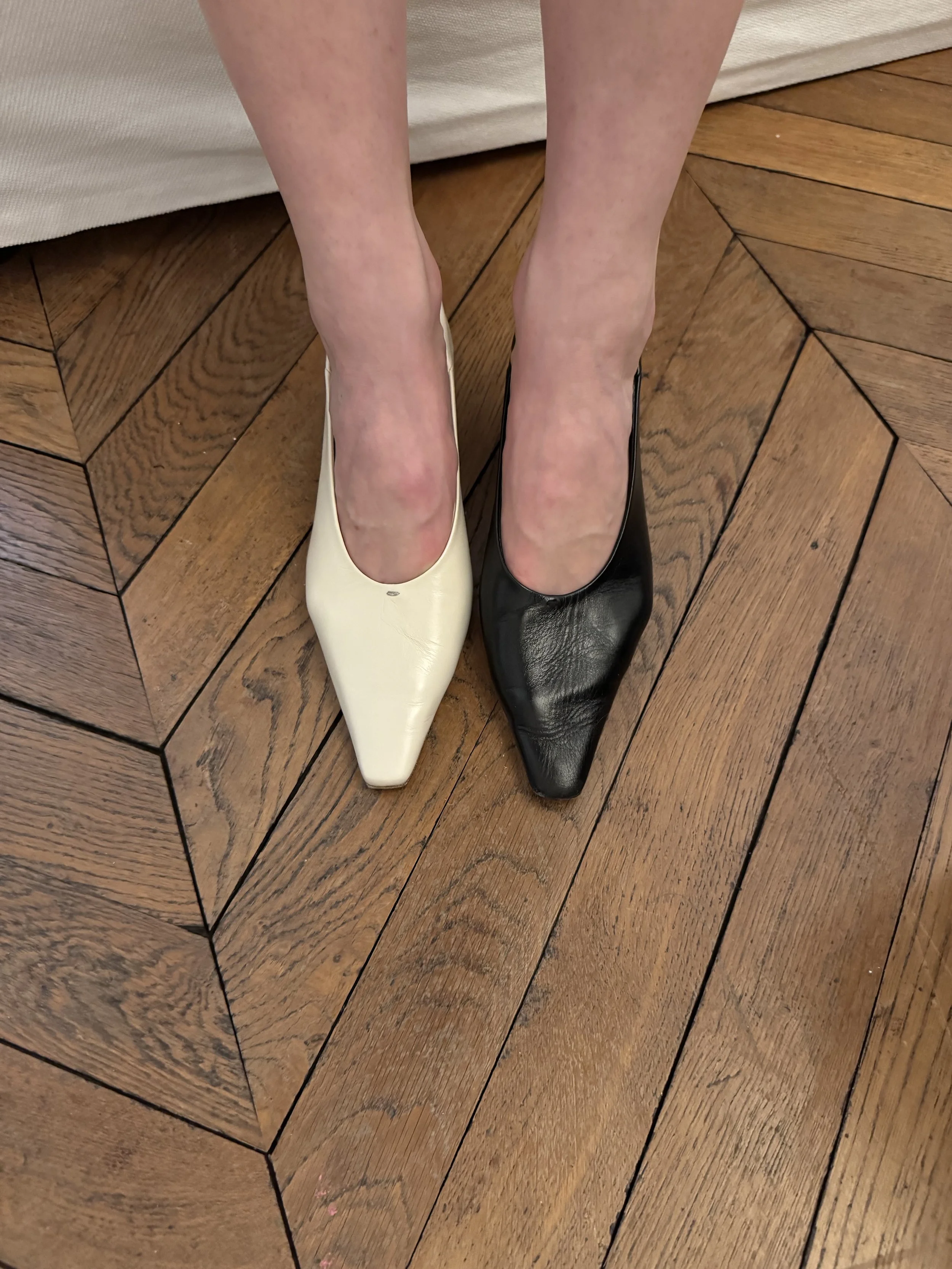 A person wearing two different color high-heeled shoes, one cream and one black, standing on a wooden floor.