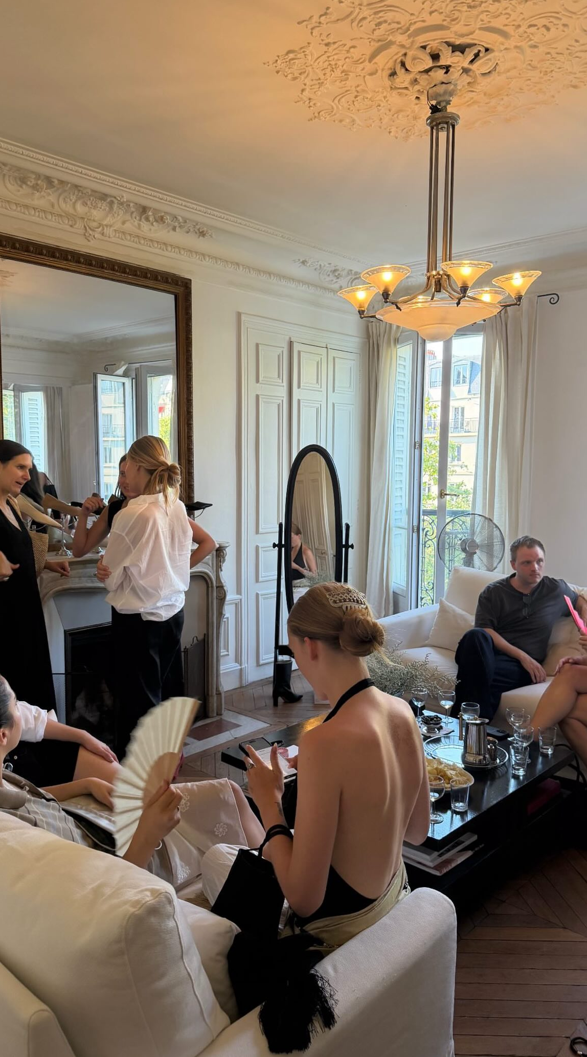 People gathered in a bright, elegant living room with white walls, large windows, and ornate ceiling molding. Some individuals are seated on a white sofa and chairs, engaging in conversation, while others stand near a fireplace and mirror. A ceiling chandelier hangs from the decorative ceiling.