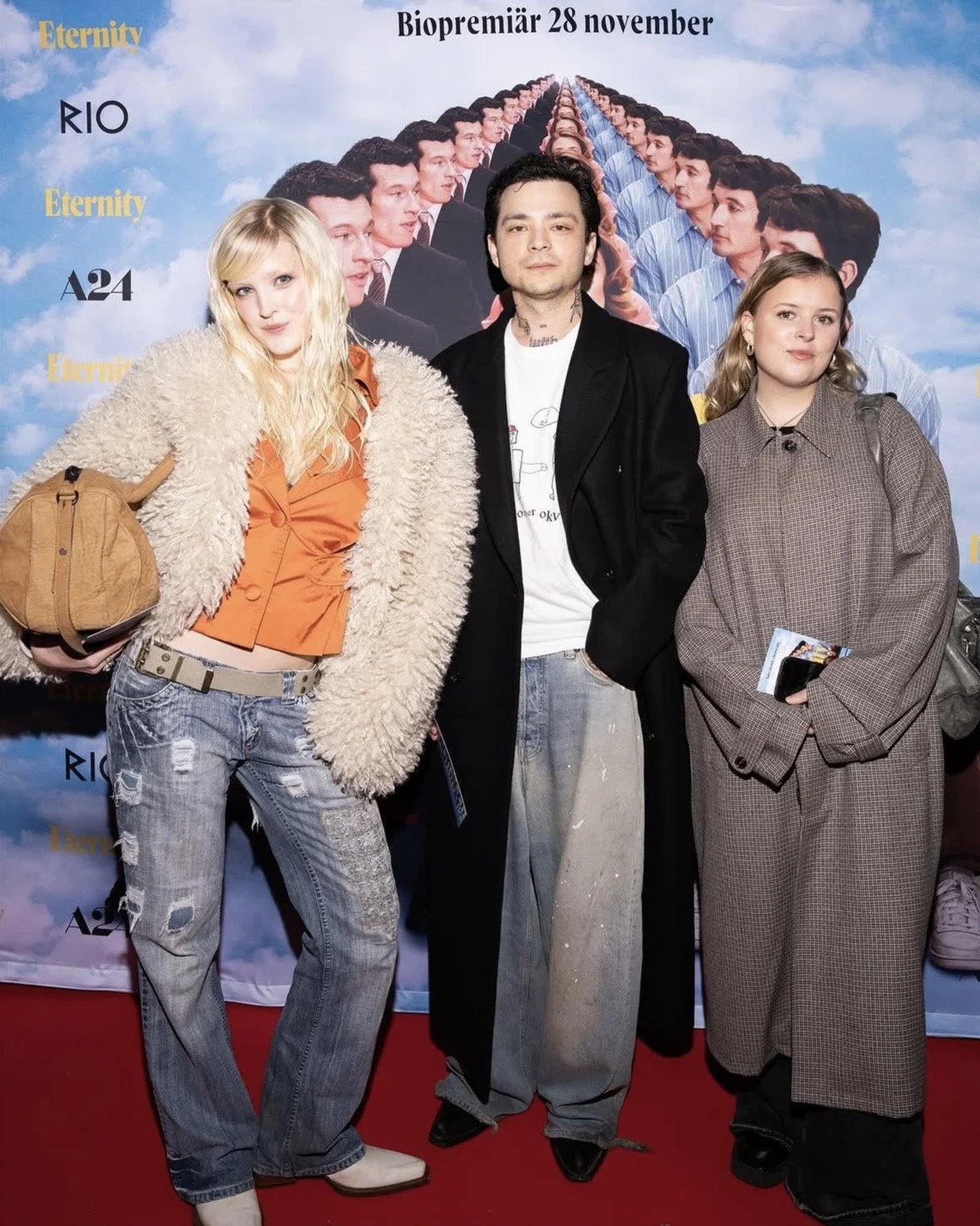 Three young people posing in front of a backdrop with a repeating image of a row of men in suits. The backdrop includes text in Swedish about a film's biographical release date, with the movie titles 'Eternity', 'RIO', and 'A24' visible.