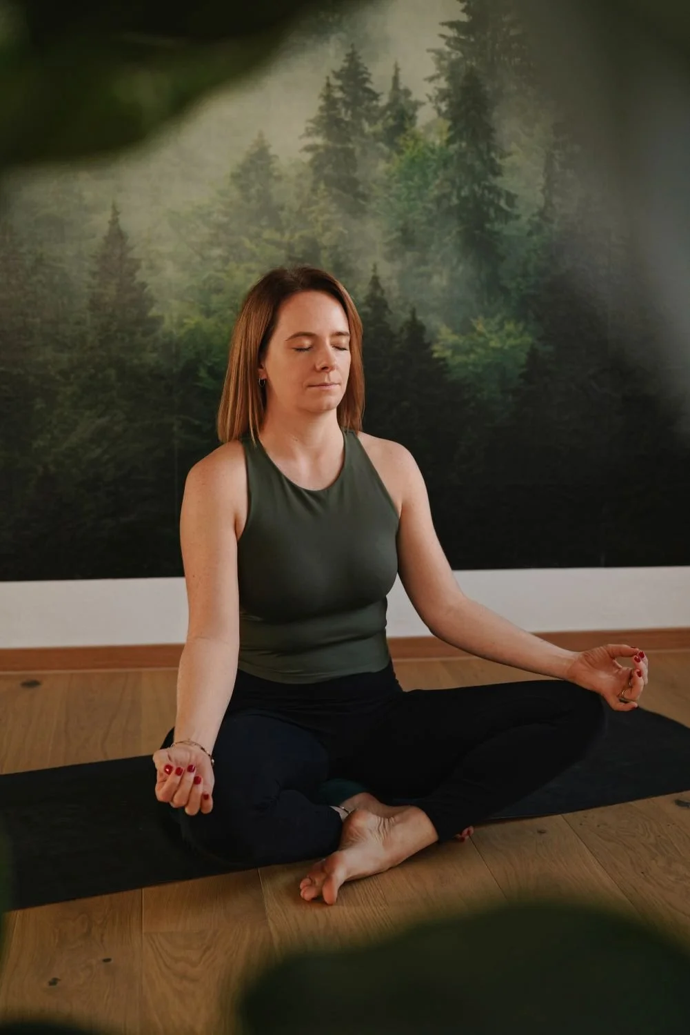 Frau in einer Yoga-Pose, sitzend im Schneidersitz, mit geschlossenen Augen in einem Raum mit Wandbild eines Waldes.