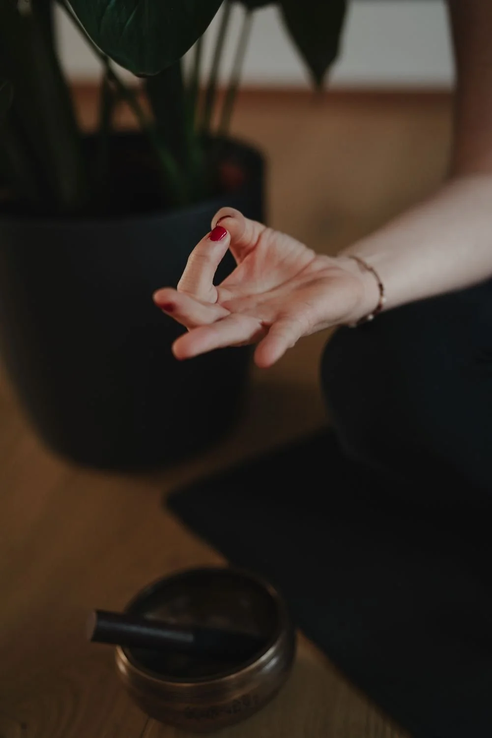 Nahaufnahme einer Hand mit roten Nagellack, die auf einem Bein im Schneidersitz ruht, neben einem schwarzen Blumentopf mit einer großen grünen Pflanze und einer Klangschale.