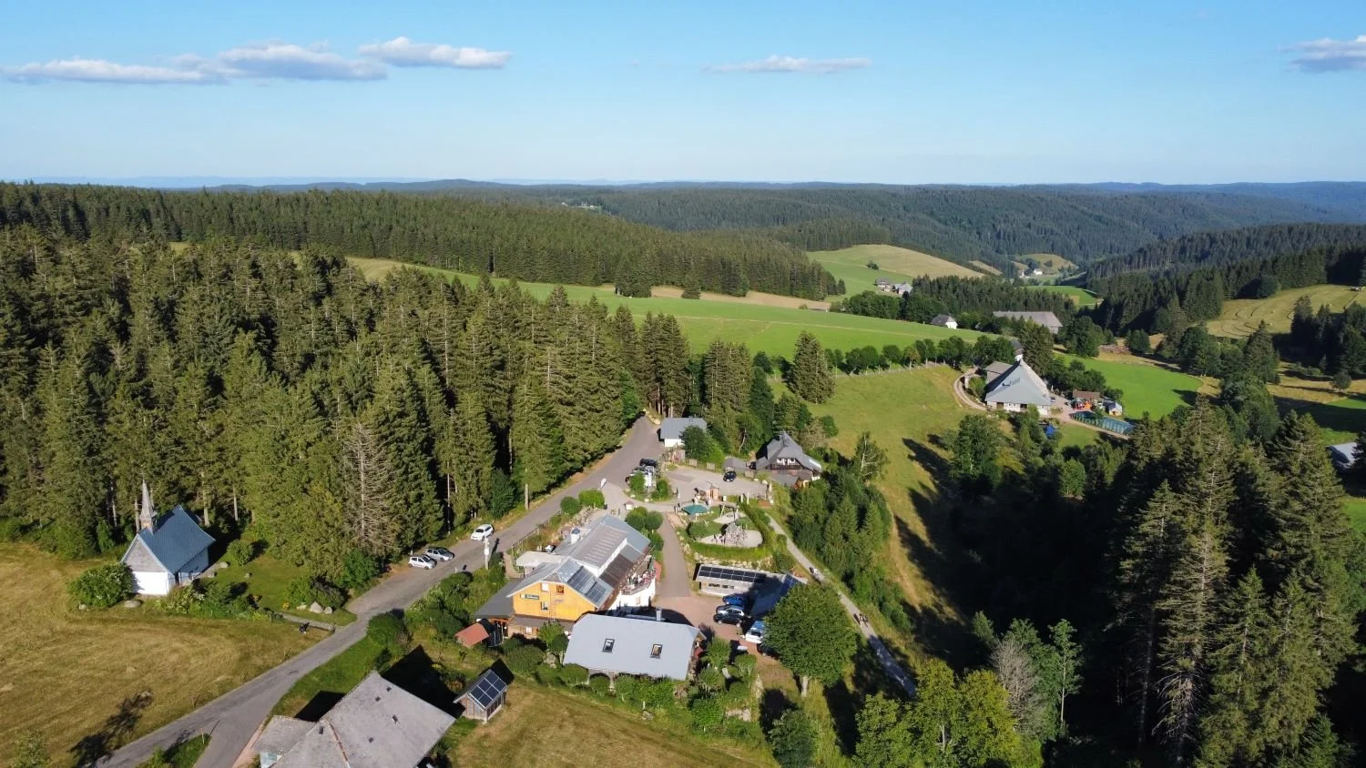 Luftaufnahme einer ländlichen Gegend mit Häusern, einer Kirche, Wäldern und Feldern bei sonnigem Himmel.
