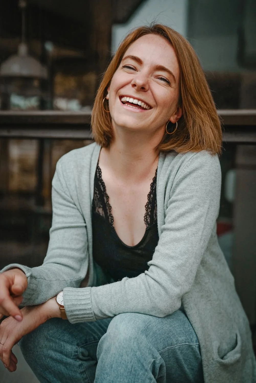 Lächelnde junge Frau mit roten Haaren, grauem Pullover, schwarzem Oberteil mit Spitze, Jeans und goldenen Ohrringen, sitzt draußen vor einem Fenster.