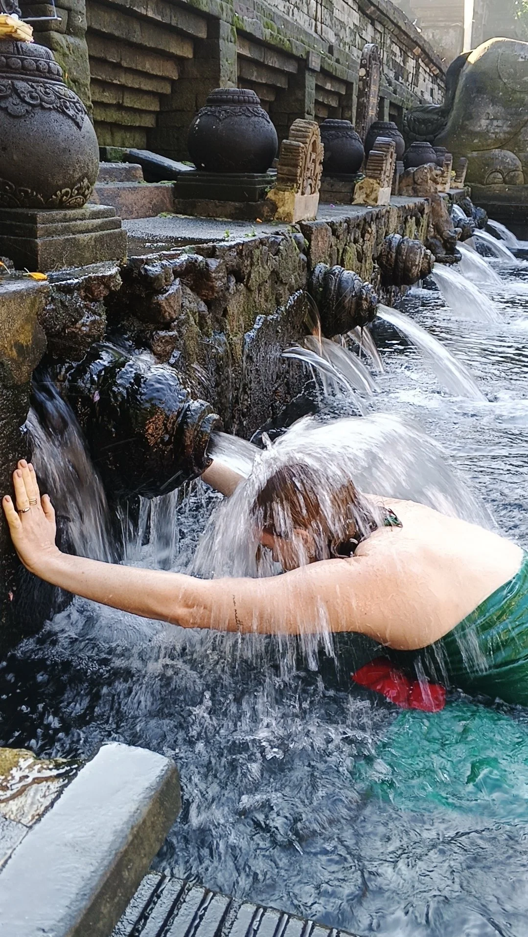 Person taucht kopfüber in einen Wasserlauf an einem mit Steinen und alten Wasserfällen geschmückten Wasserweg, während Wasser über die Wasserrohre fließt.