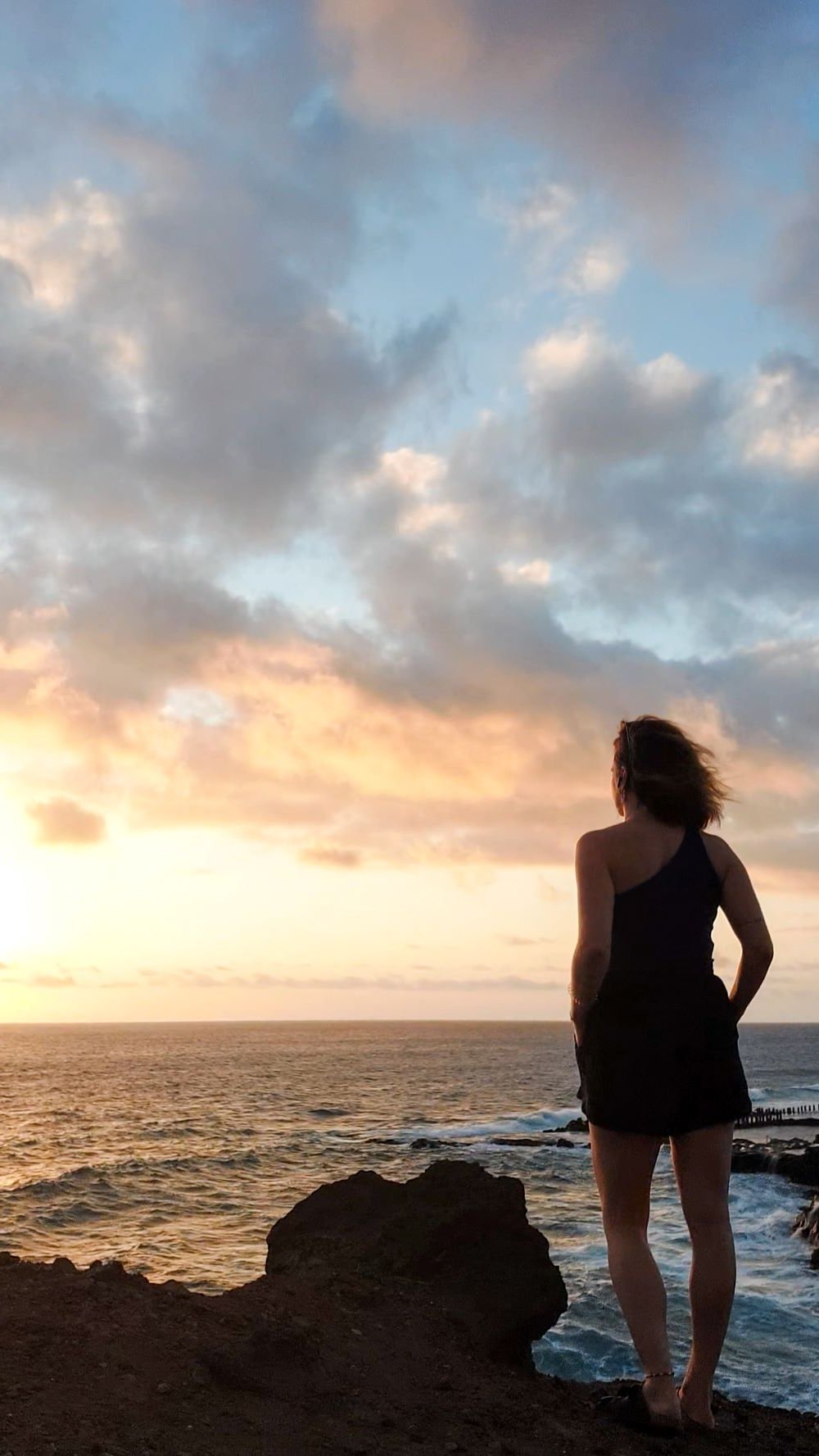 Eine Frau in einem schwarzen Kleid steht an einer felsigen Küste und blickt auf das Meer bei Sonnenuntergang, mit einem dramatischen Himmel voller Wolken darüber.