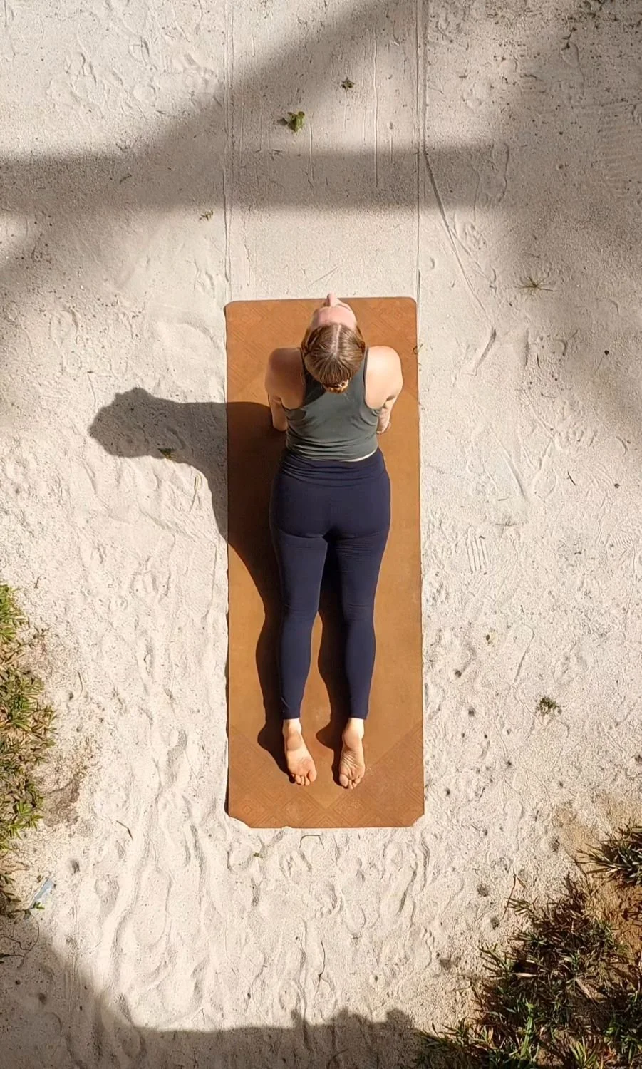 Eine Frau macht Yoga auf einer Yoga-Matte im Freien auf sandigem Boden, aufgenommen von oben.