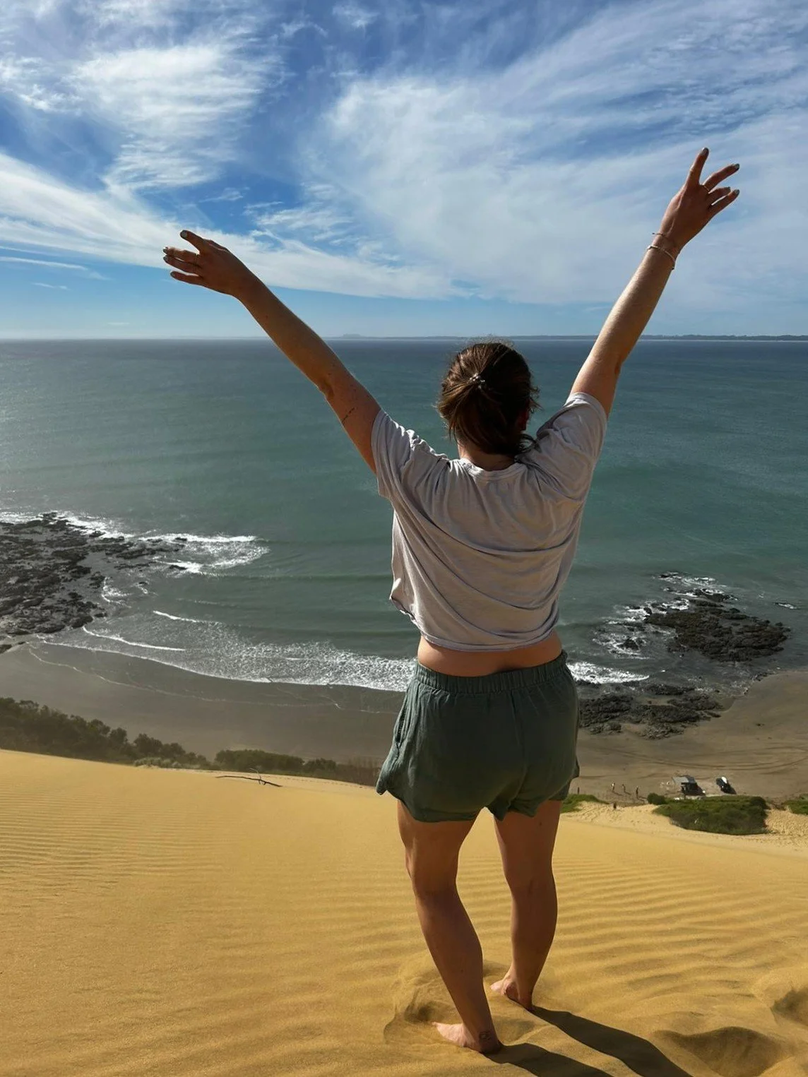 Frau steht auf einer Sanddüne am Meer und hebt die Arme in die Luft bei Sonnenuntergang oder -aufgang.