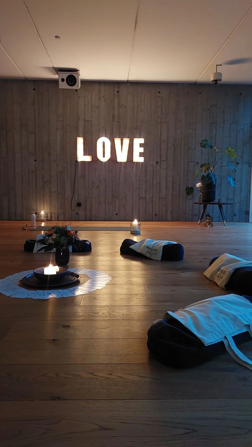Ein Raum mit Holzboden und einer Holzvertäfelung an der Wand. Auf der Wand steht eine leuchtende Dekoration mit dem Wort "LOVE". Im Raum sind Kerzen, ein Blumenarrangement, gepäckartige Gegenstände mit weißen Tüchern und eine Pflanze auf einem kleinen Tisch.