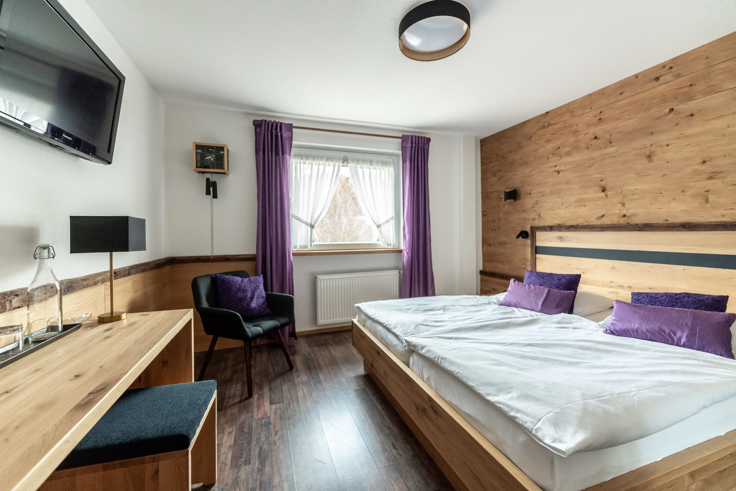 Ein gemütliches Schlafzimmer mit Holzdeckenwand, weißen Wänden, einem Fenster mit lila Vorhängen, einem Bett mit lila Kissen, einem schwarzen Sessel, einem Holzschreibtisch mit schwarzer Lampe, einer Wanduhr, einem Fernseher an der Wand und einem Radiator unter dem Fenster.