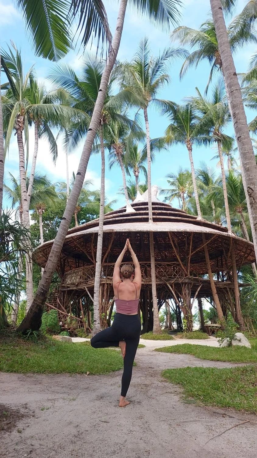 Frau praktiziert Yoga im Freien vor einem runden, strohgedeckten Hütte, umgeben von hohen Palmen auf einer tropischen Insel.