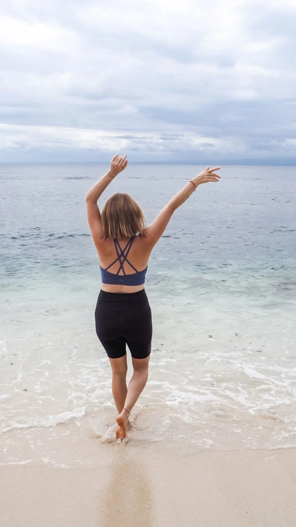 Eine Frau steht am Strand mit Blick aufs Meer, hebt die Arme in die Luft und genießt die Natur.