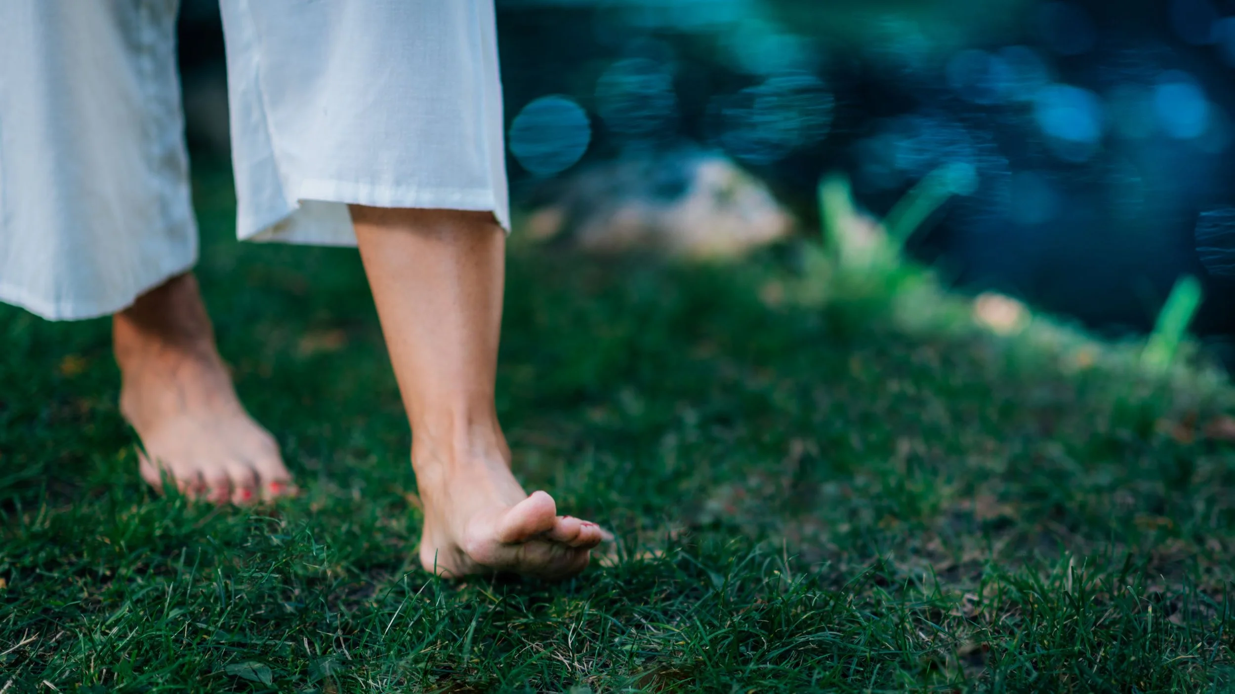 Füße einer Person, die barfuß auf Gras im Freien steht, mit weißen Hosen und einem fließenden weißen Oberteil, bei Sonnenlicht.