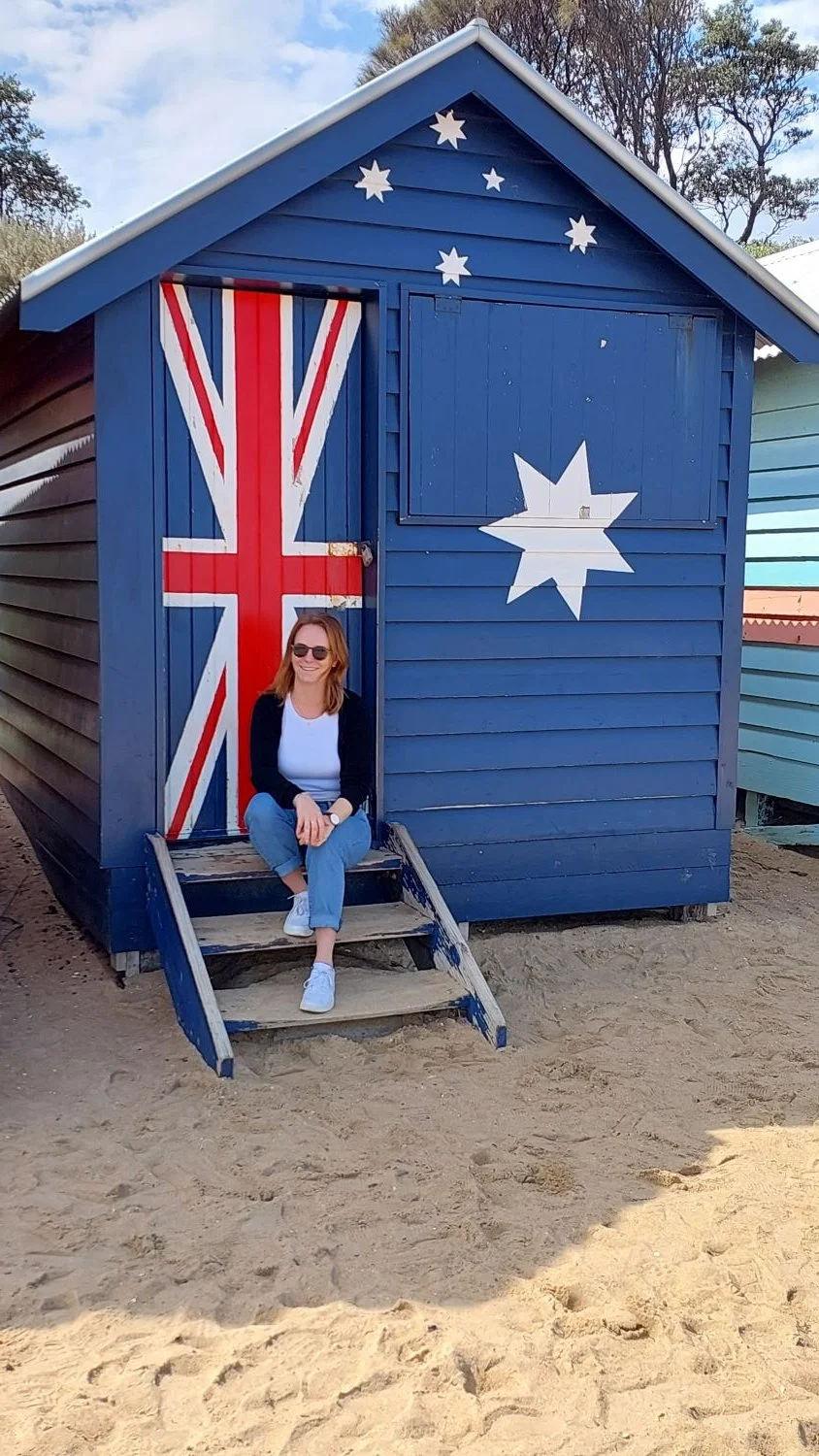 Eine Frau sitzt auf den Treppen eines blauen Strandhauses, das mit dem Union Jack (britische Flagge) und australischen Sternen bemalt ist. Das Haus hat eine blaue Fassade mit weißen Sternen und Flaggenmuster auf der Tür.
