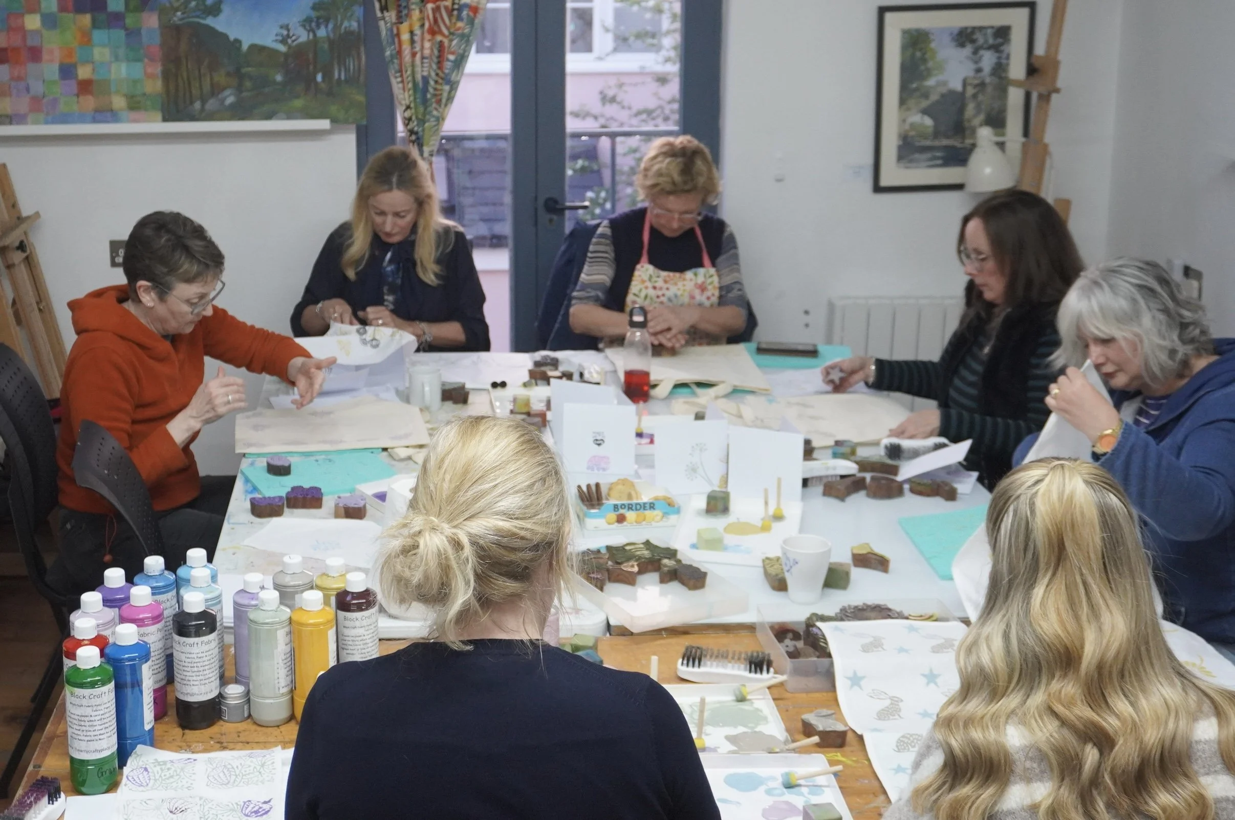 A group of women is sitting around a table engaging in a craft activity, working with paints and various craft supplies. The table is covered with paper, paint bottles, and decorated blocks. The room has colorful artwork on the walls and a glass door leading outside.