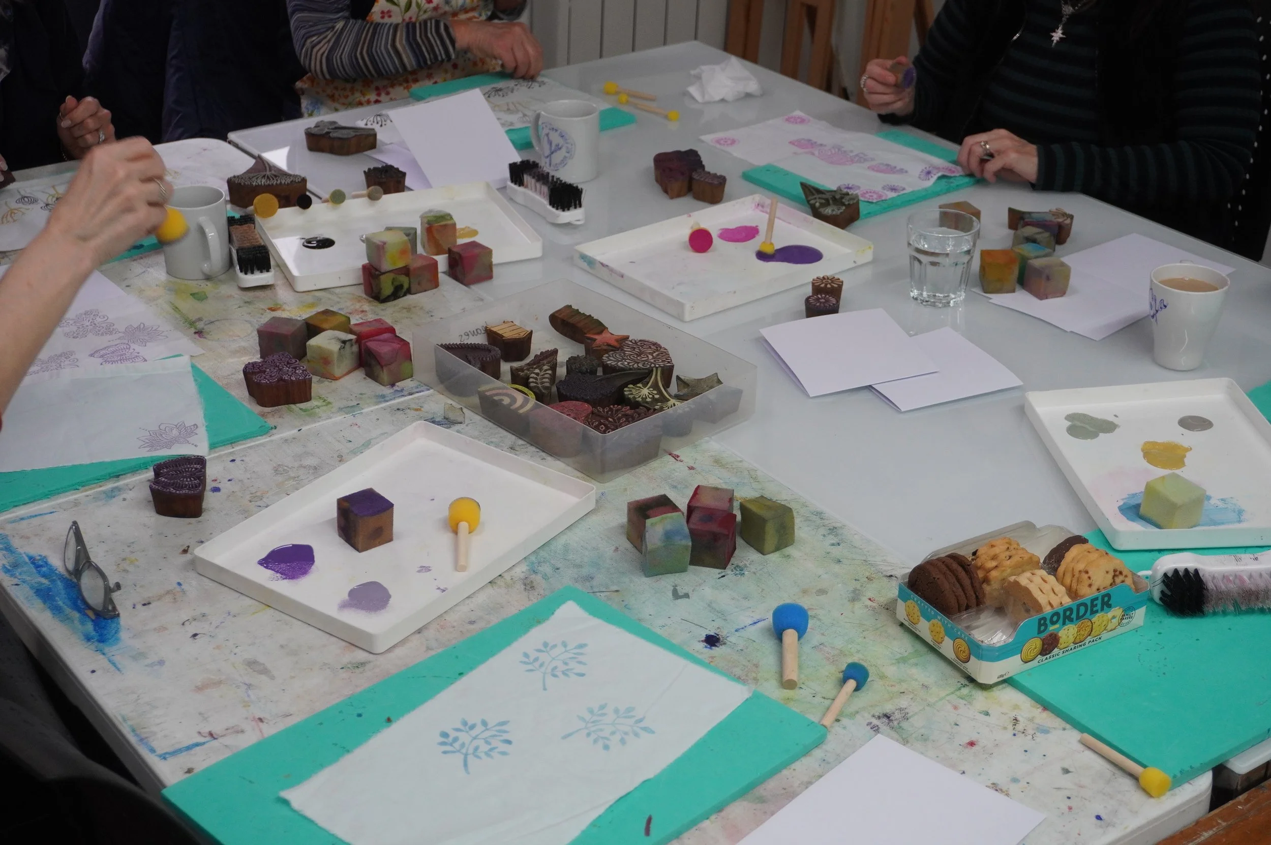 A table with various handmade soap pieces of different colors and shapes, some with intricate designs, along with soap-making tools, tissue papers, cups, and snacks, indicating a soap-making workshop or class.