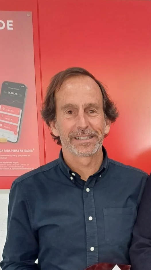 Portrait of a middle-aged man with brown hair and a beard, wearing a navy blue button-up shirt, standing in front of a red background with a poster and a smartphone image.