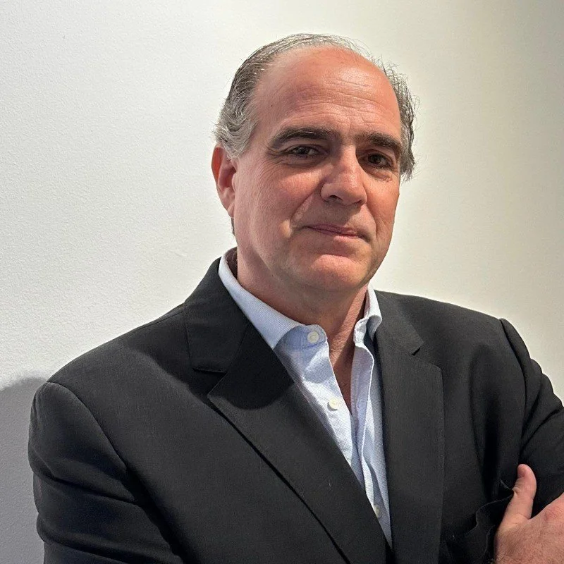 A middle-aged man in a black suit and white dress shirt standing against a plain white wall, with his arms crossed and a confident expression.