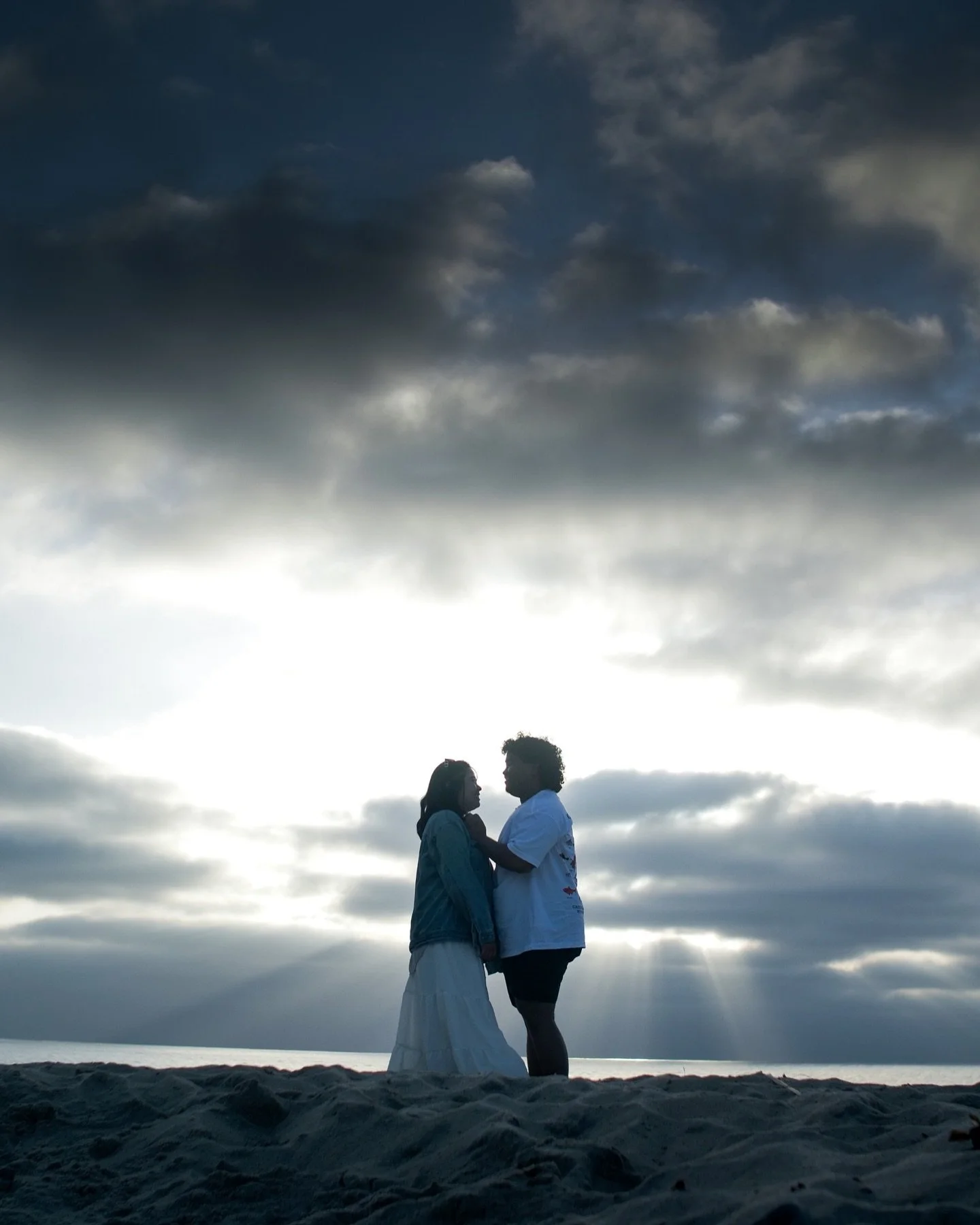 A couple standing close together on a beach at sunset, with dark clouds overhead and the sun shining behind them.