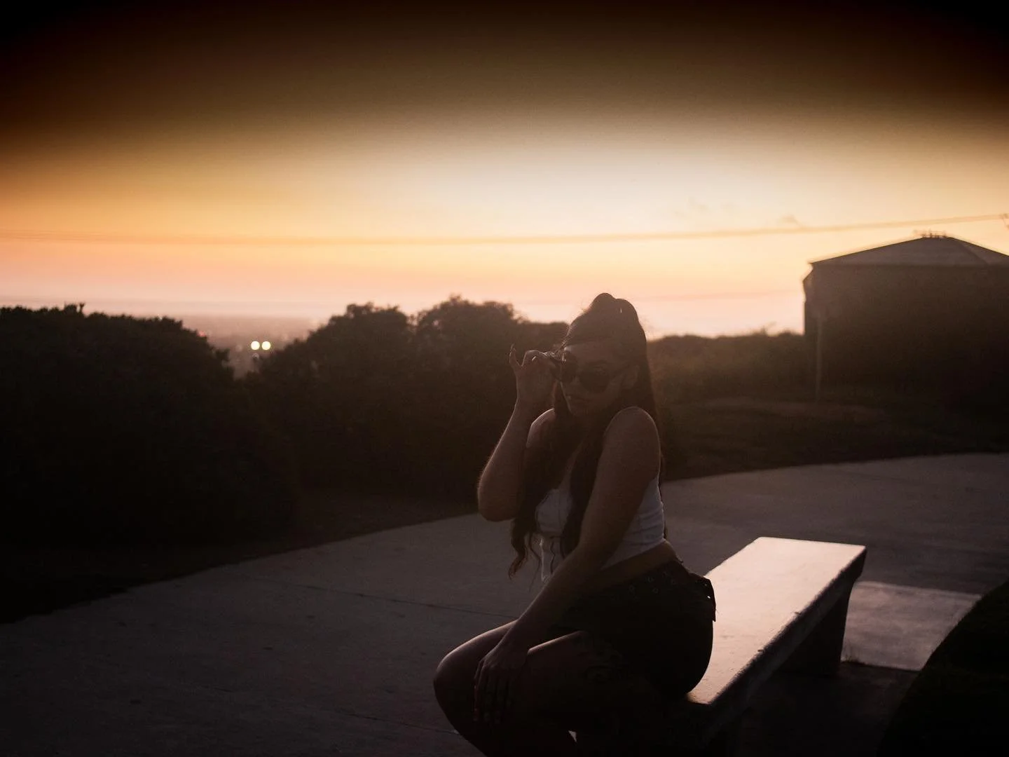 Silhouette of a woman with long hair, sunglasses, and a white tank top sitting on a bench at sunset, with trees and a building in the background.