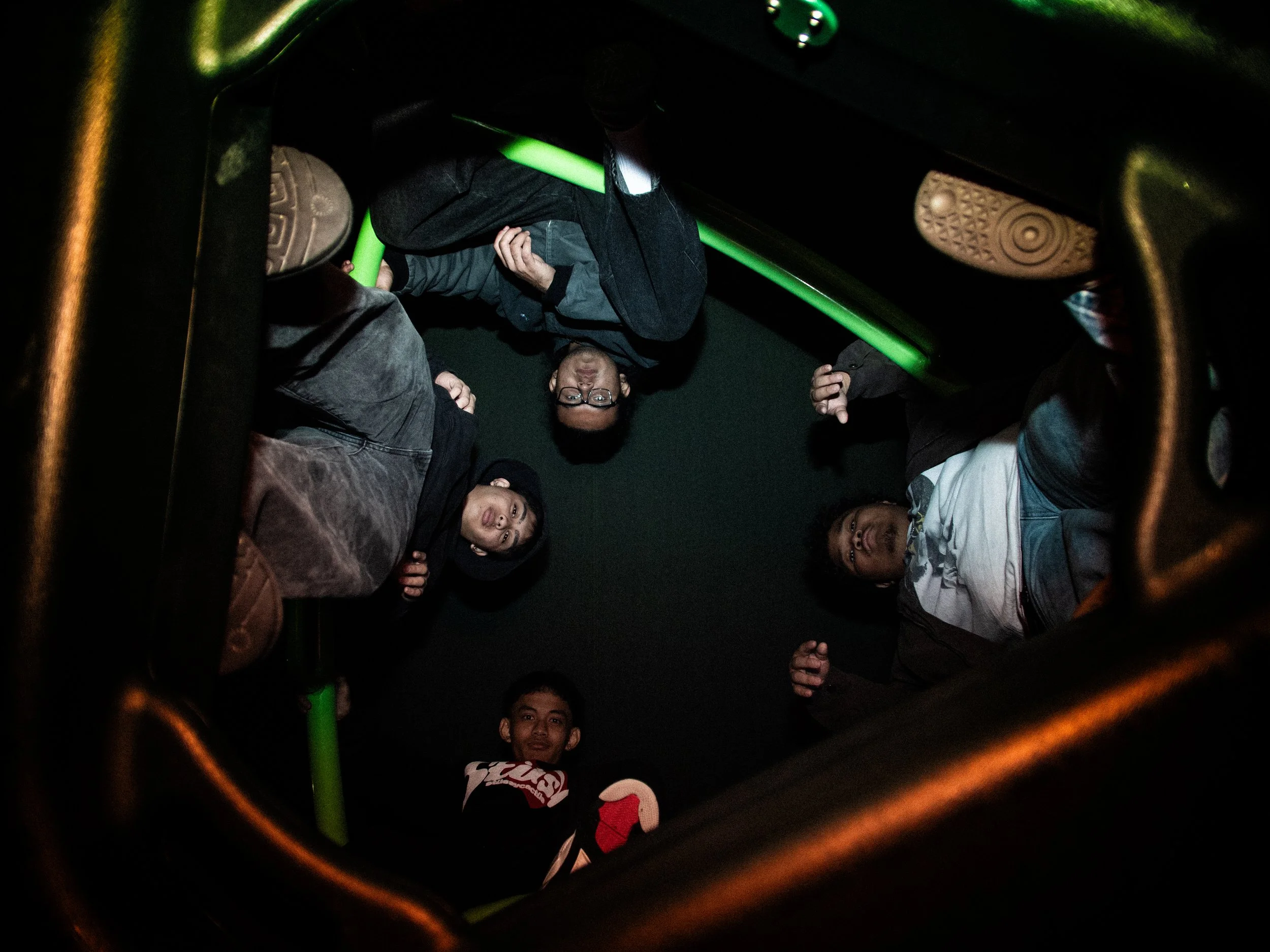 A group of young men standing inside what appears to be a dark room or inside a vehicle, looking down toward the camera. The photo is taken from below, with the image framed by orange and green handlebars or bars around the edges.