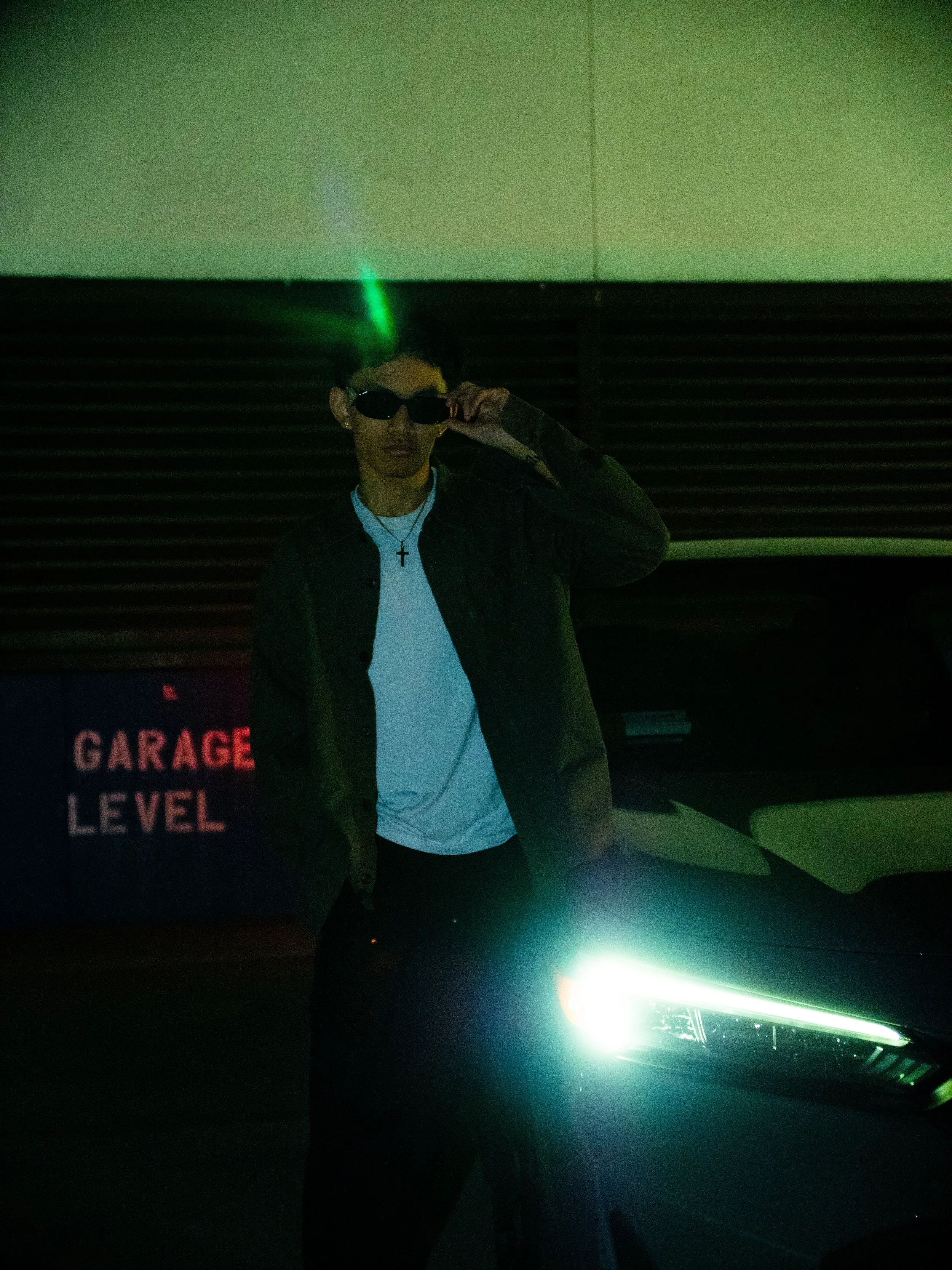 Person wearing sunglasses and a necklace with a cross, standing in front of a car with bright headlights on, in a dark garage with a sign reading 'GARAGE LEVEL' in the background.