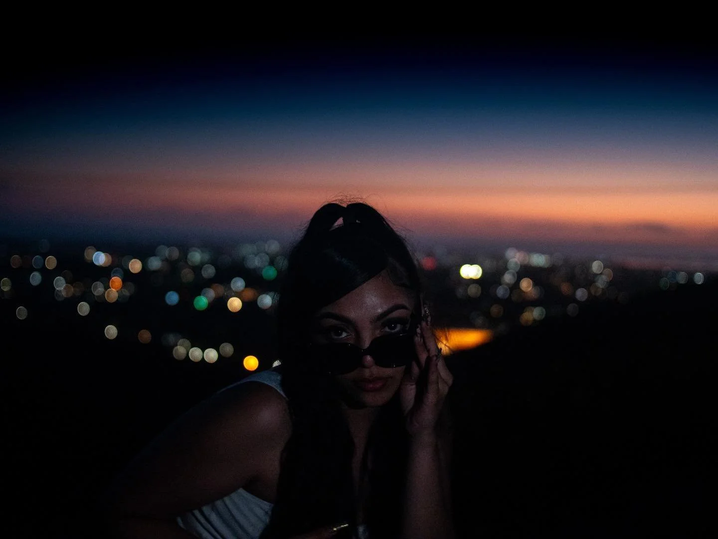 A woman wearing sunglasses and a white top, posing with her hand near her face, against a colorful sunset sky and city lights in the distance.