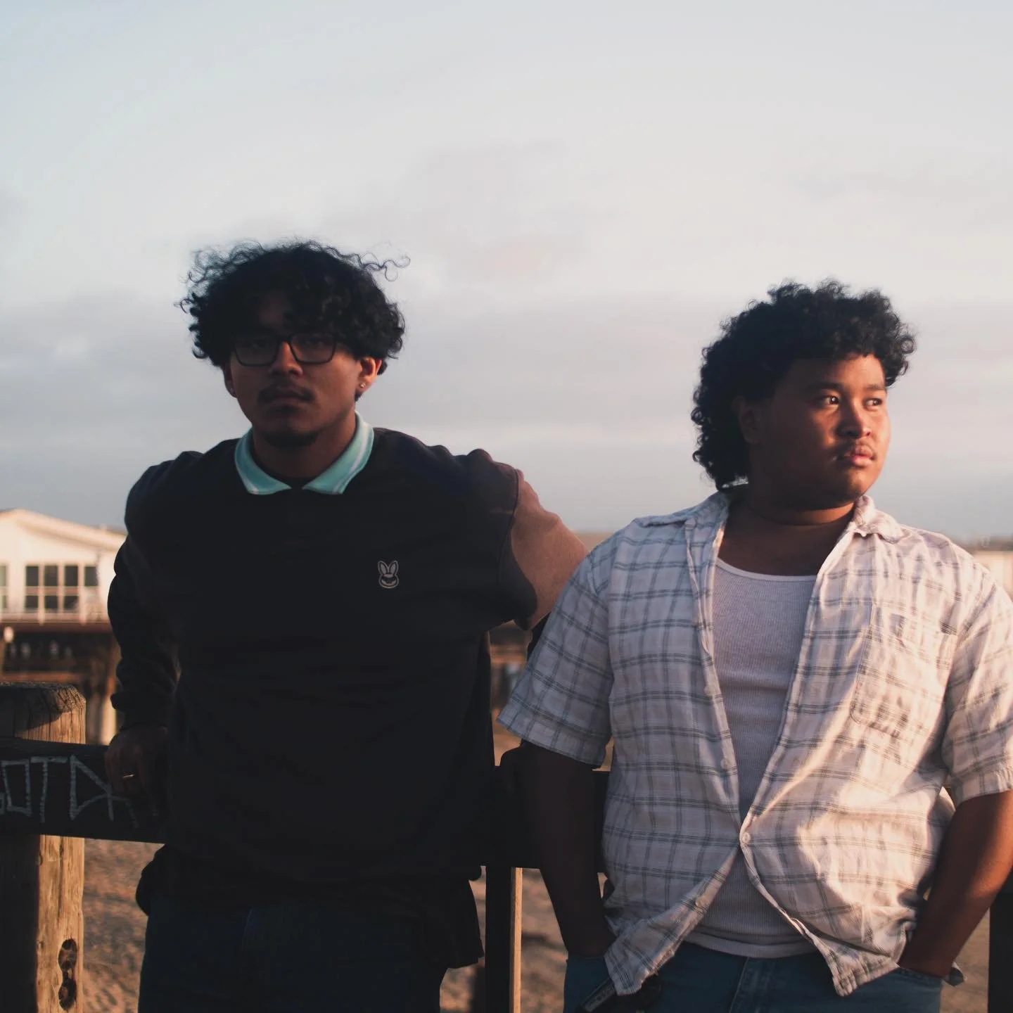 Two young men with curly hair standing outdoors at sunset, one in a black shirt with glasses and the other in a light checkered shirt, near a wooden railing.