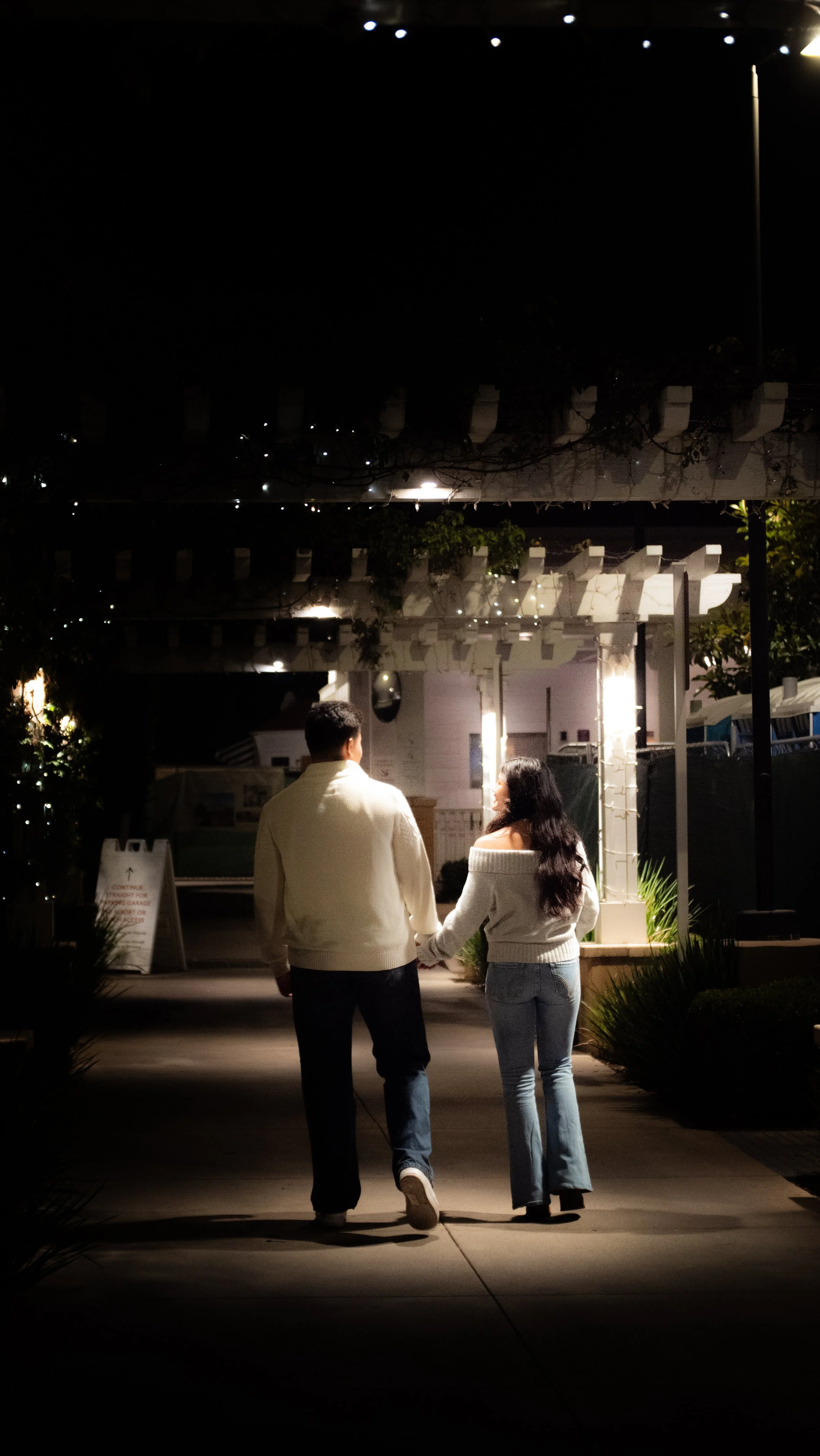 A couple holding hands walking at night under string lights and a streetlamp near a building.