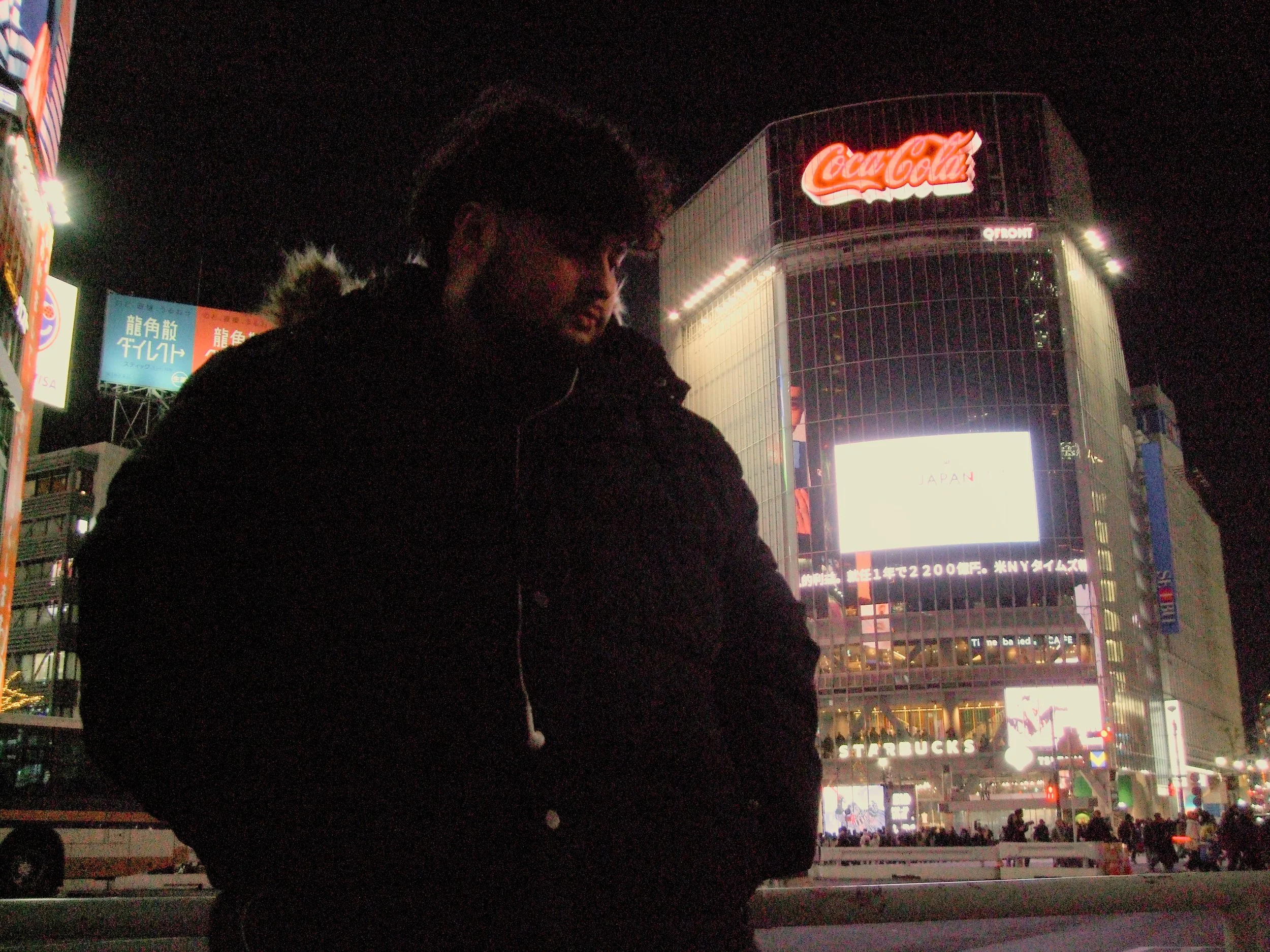 Nighttime scene in Tokyo, Japan with a person in the foreground and a brightly lit building with a Coca-Cola sign in the background.