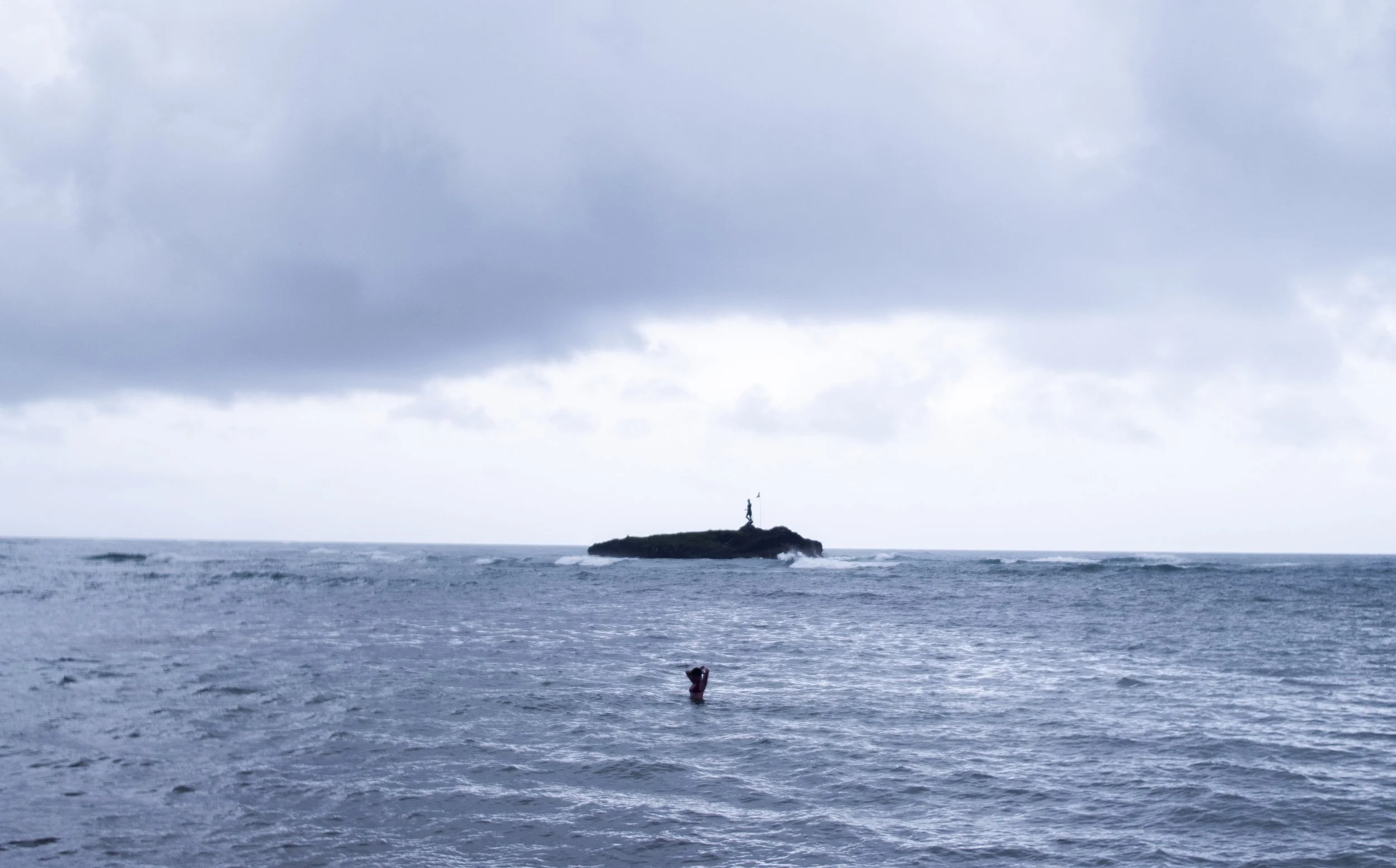 A lone person standing in the ocean near a small rock island with a flag, with cloudy sky overhead.