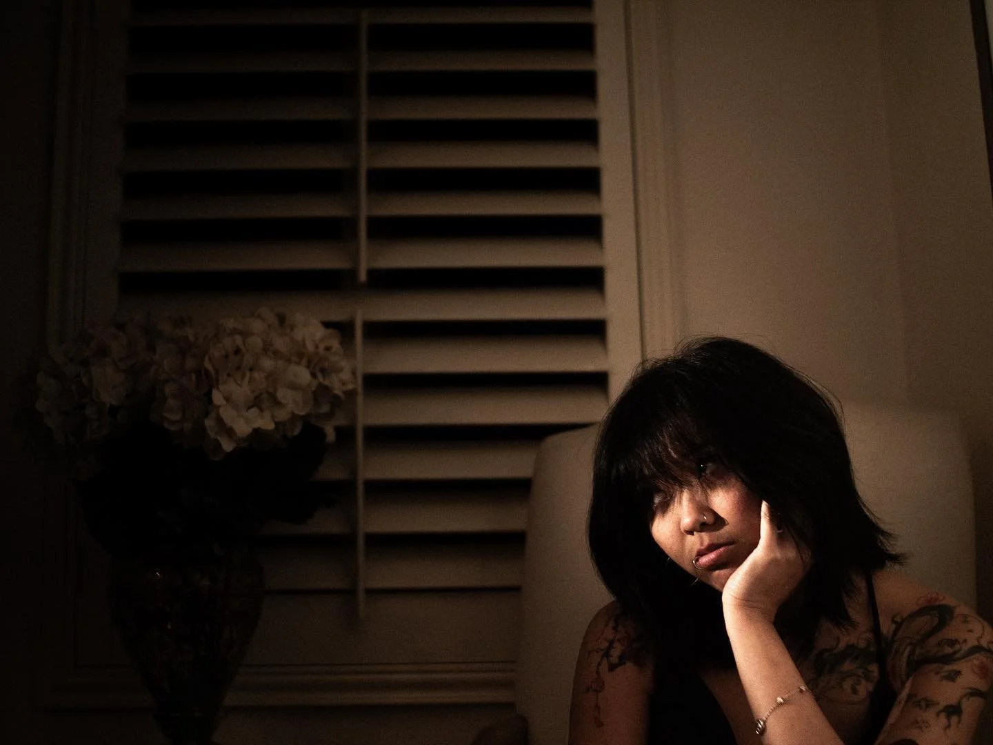 A woman with black hair and tattoos, resting her head on her hand, sitting on a couch in a dimly lit room.