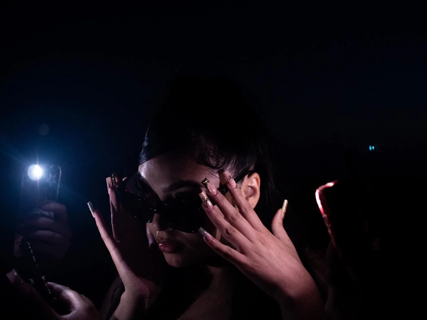 Woman with long dark hair and sunglasses covering her eyes, holding a phone with a light in a dark environment.