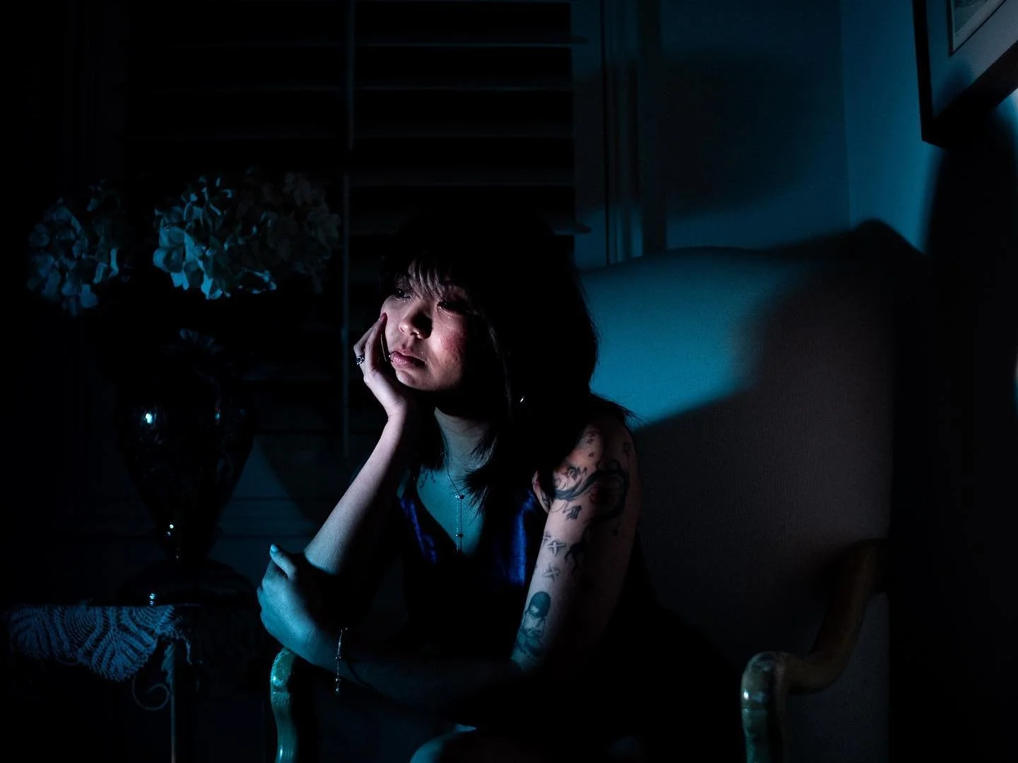 A young woman with tattoos and dark hair sits in a dimly lit room, resting her head on her hand, looking thoughtful or pensive.