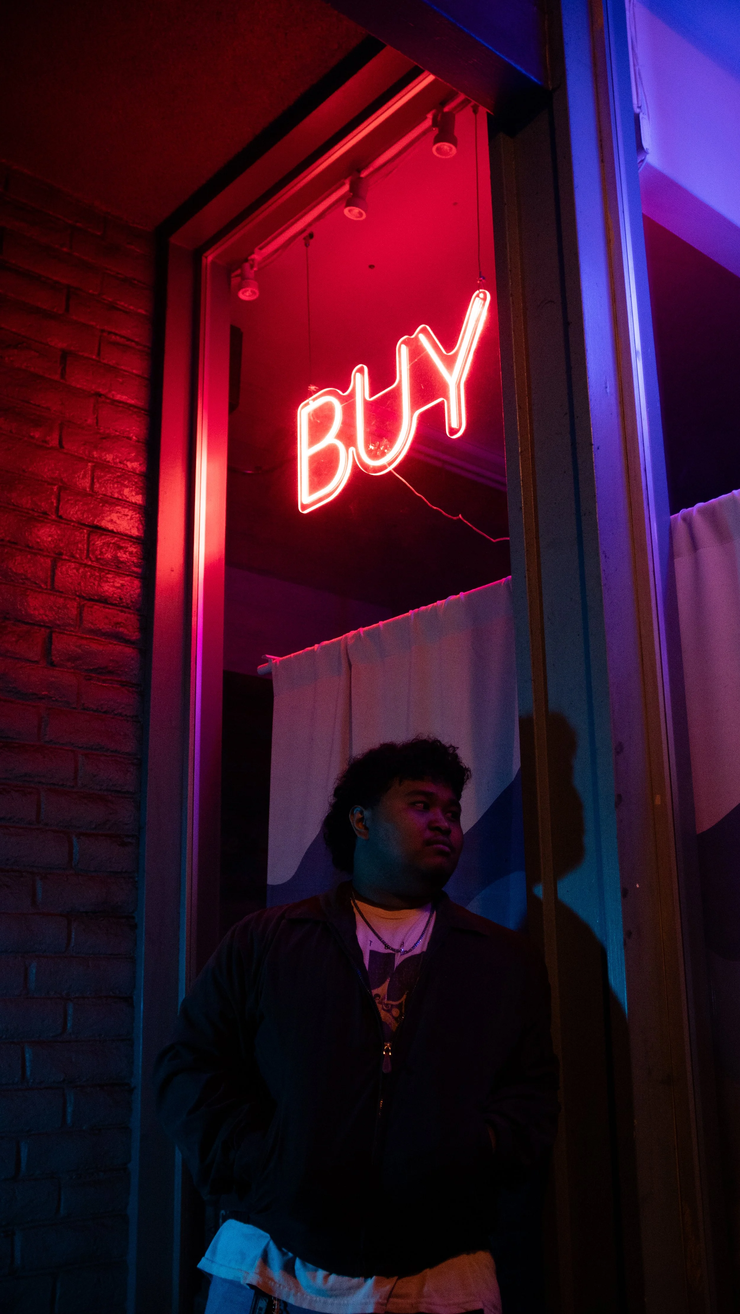 Neon sign displaying the word 'BUY' in red above a person with curly hair, wearing a dark jacket, standing outside a building with brick and wooden wall sections at night.