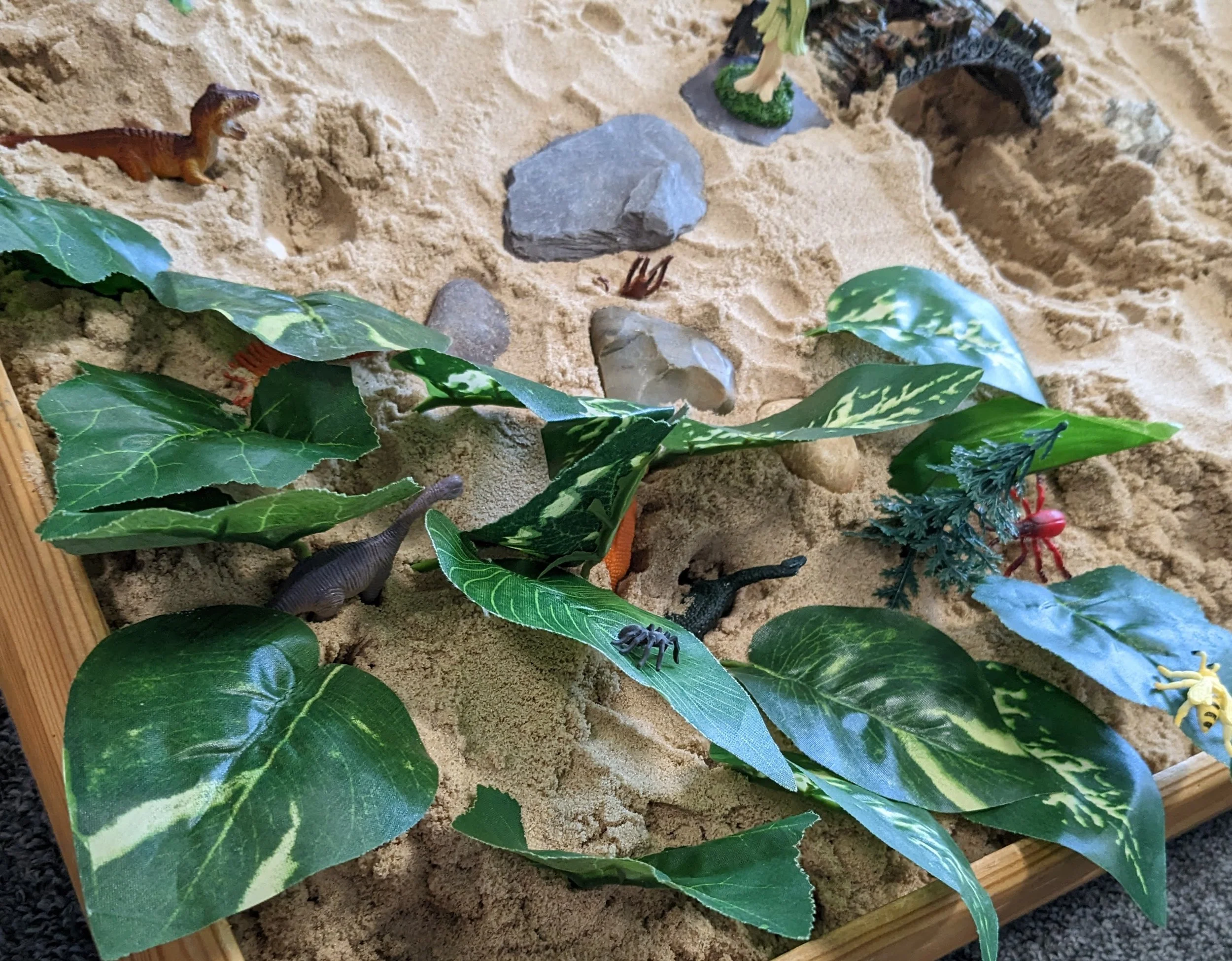 A close-up of a sandbox with plastic dinosaur and crab figurines, green plants, and rocks.