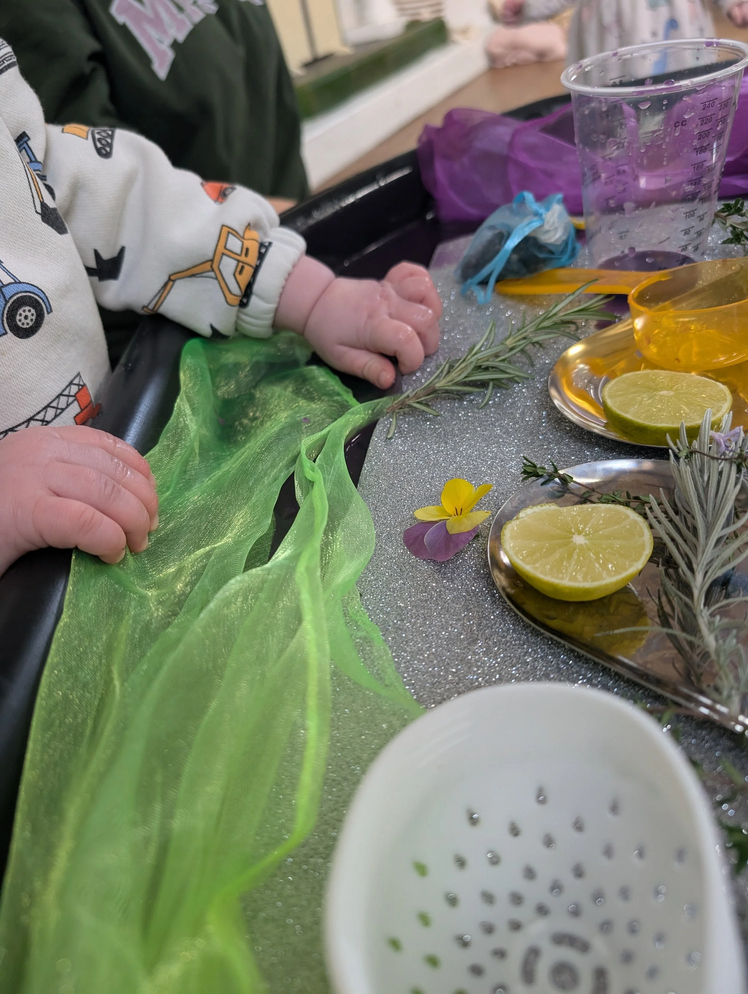 Close-up of a baby's hand resting on a green ribbon or fabric, surrounded by cups, herbs, lemon slices, and flowers on a table.