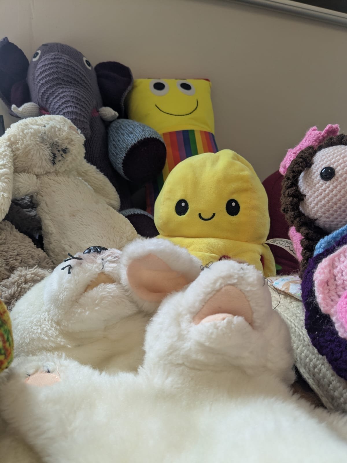 A collection of plush toys including a cream-colored sheep, a gray elephant, a yellow smiling stuffed character with black eyes and striped rainbow torso, and a crocheted doll with dark hair and pink bow, surrounded by various other stuffed animals.